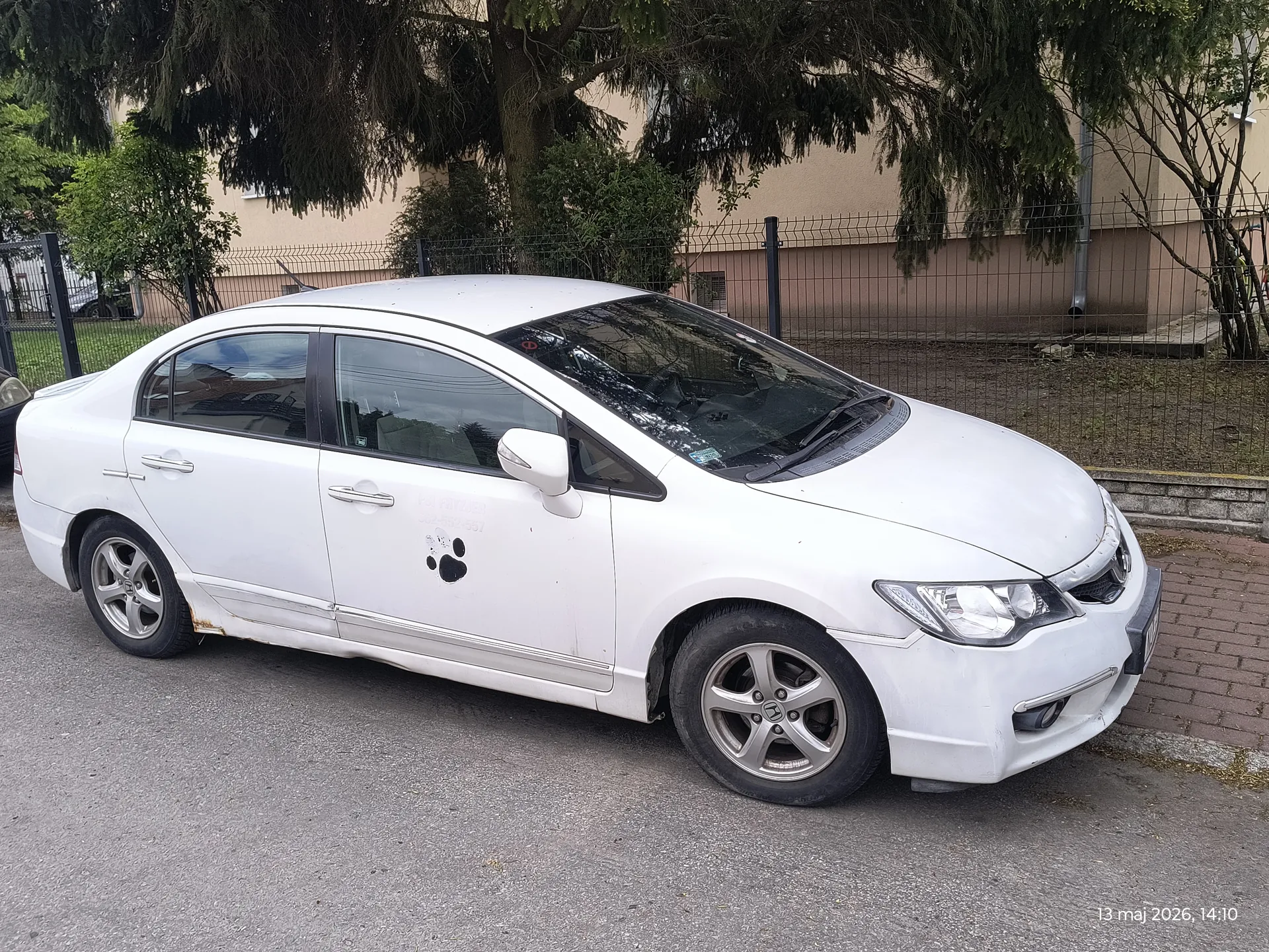 Biały samochód osobowy typu sedan zaparkowany na asfaltowej nawierzchni. Na drzwiach pasażerskich widoczny czarny odcisk łapy. Samochód posiada srebrne felgi, a na przednim zderzaku oraz nadkolu widoczne ślady korozji. W tle metalowe ogrodzenie, drzewa, krzewy oraz beżowy budynek. W prawym dolnym rogu obrazu znajduje się data i czas: 13 maj 2026, 14:10.