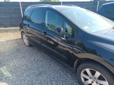 Czarny samochód typu kombi zaparkowany na żwirowym podłożu na tle siatkowego ogrodzenia. Widoczny jest prawy bok pojazdu, w tym przednie i tylne drzwi, lusterko boczne oraz felgi aluminiowe.