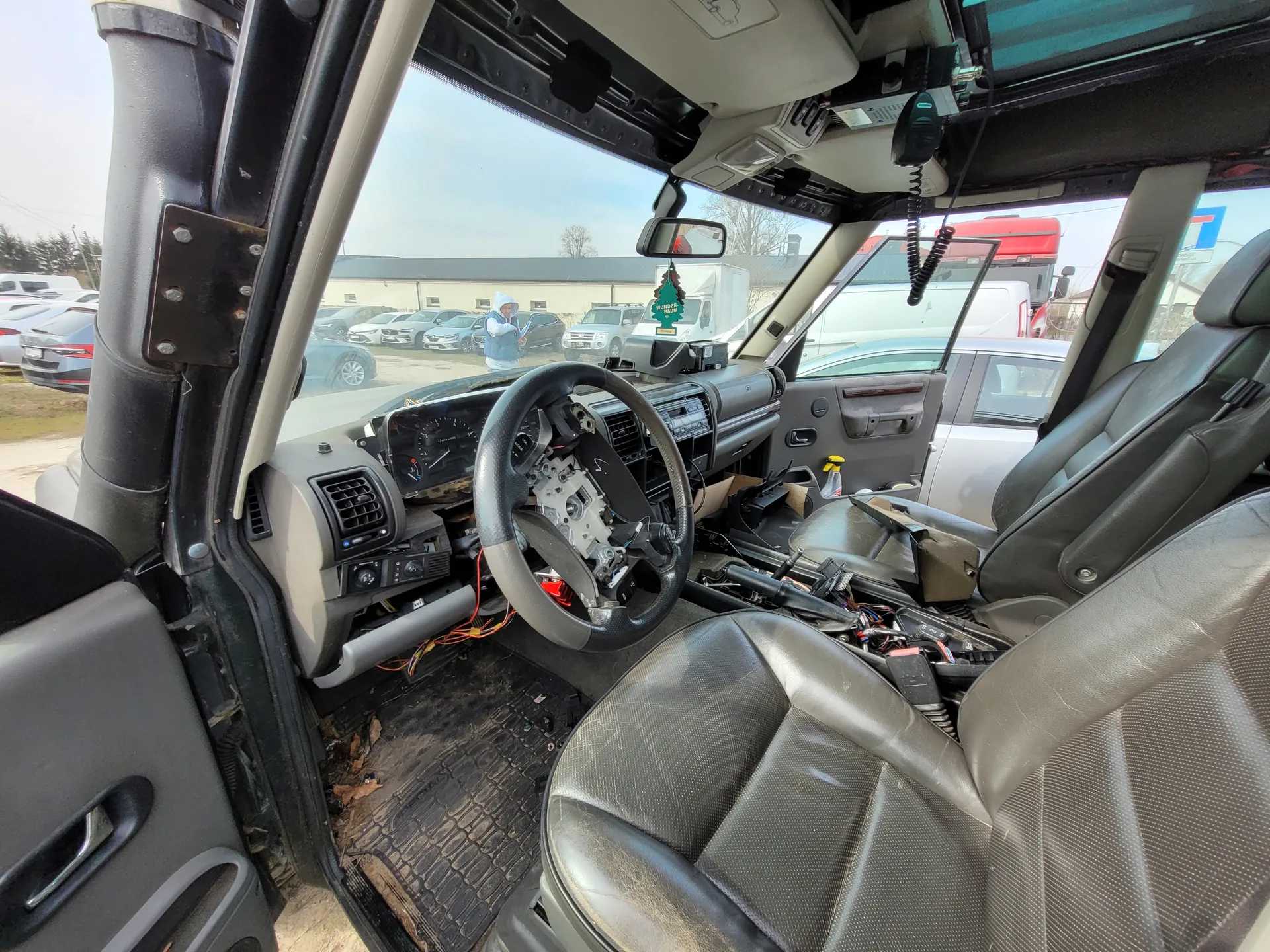 Wnętrze samochodu z rozebraną deską rozdzielczą, odsłoniętymi przewodami i częściowo demontowanym kierownicą. Widoczne są czarne skórzane fotele, otwarte drzwi po stronie kierowcy oraz zewnętrzny parking z innymi pojazdami. Na lusterku wstecznym wisząca ozdoba w kształcie choinki. W tle budynki i osoba stojąca na zewnątrz.