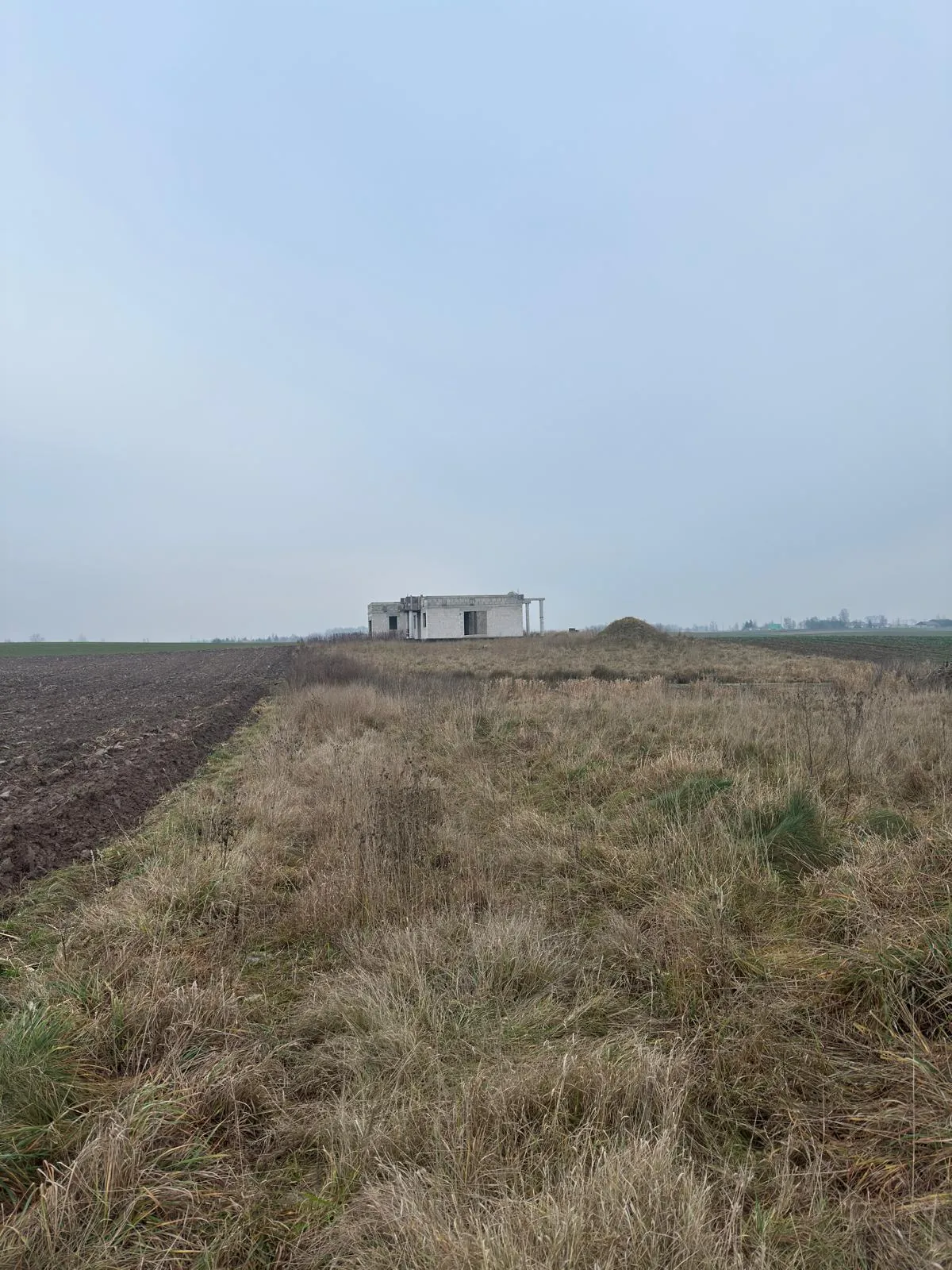 Pole z suchą trawą na pierwszym planie, po lewej stronie uprawa z ornym polem. W oddali znajduje się niedokończony budynek z betonowych bloków z otwartymi przestrzeniami zamiast drzwi i okien. Niebo jest jasne, bezchmurne. Linia horyzontu jest pozioma, a całość ujęcia jest stabilna.