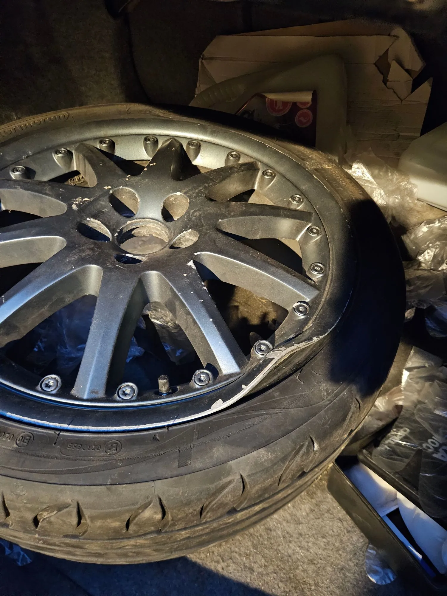 Zbliżenie na srebrną, wieloramienną felgę aluminiową z oponą samochodową. Felga posiada widoczne uszkodzenia mechaniczne w postaci wgniecenia i zarysowań na zewnętrznym rancie. Koło leży poziomo na podłożu w ciemnym pomieszczeniu, w tle widoczne są kartony i folia plastikowa.