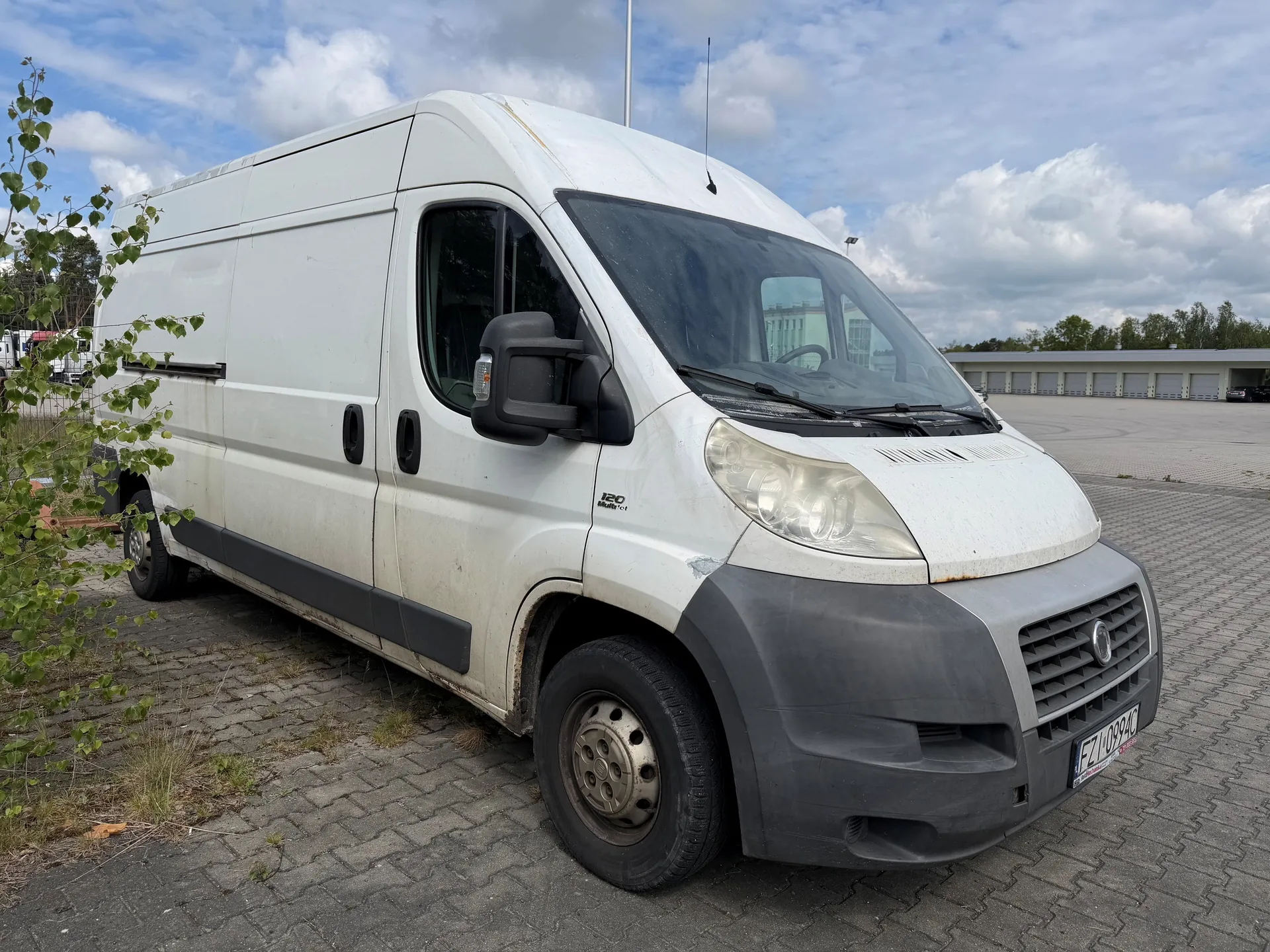 Biały furgon marki Fiat Ducato zaparkowany na brukowanym placu. Furgon ma szary przód, widoczne są reflektory, tablica rejestracyjna FZL 0994C oraz napisy 120 i Multijet na boku. W tle budynki z bramami garażowymi, drzewa i niebo z chmurami. Po lewej stronie obrazu rośnie krzew z zielonymi liśćmi. Furgon wykazuje ślady zużycia i zabrudzeń.