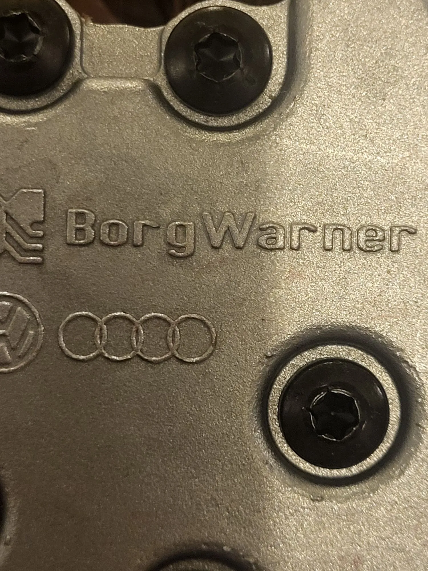 Zbliżenie na metalową, srebrną powierzchnię elementu mechanicznego. Na powierzchni znajduje się wytłoczony napis „BorgWarner” oraz logo marki Audi składające się z czterech przeplatających się pierścieni. Widoczne są również trzy czarne śruby z łbami imbusowymi.