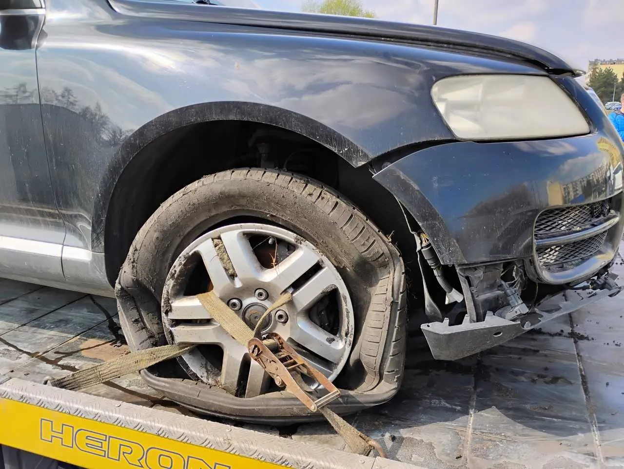 Zdjęcie przedstawia uszkodzony czarny samochód osobowy na platformie holownika. Widoczna jest przód lewej strony pojazdu z pękniętym przednim zderzakiem, uszkodzonym błotnikiem i płaską oponą przymocowaną pasami do platformy. Na żółtej części holownika widnieje napis HERON. W tle znajdują się budynki i osoba w niebieskiej odzieży.