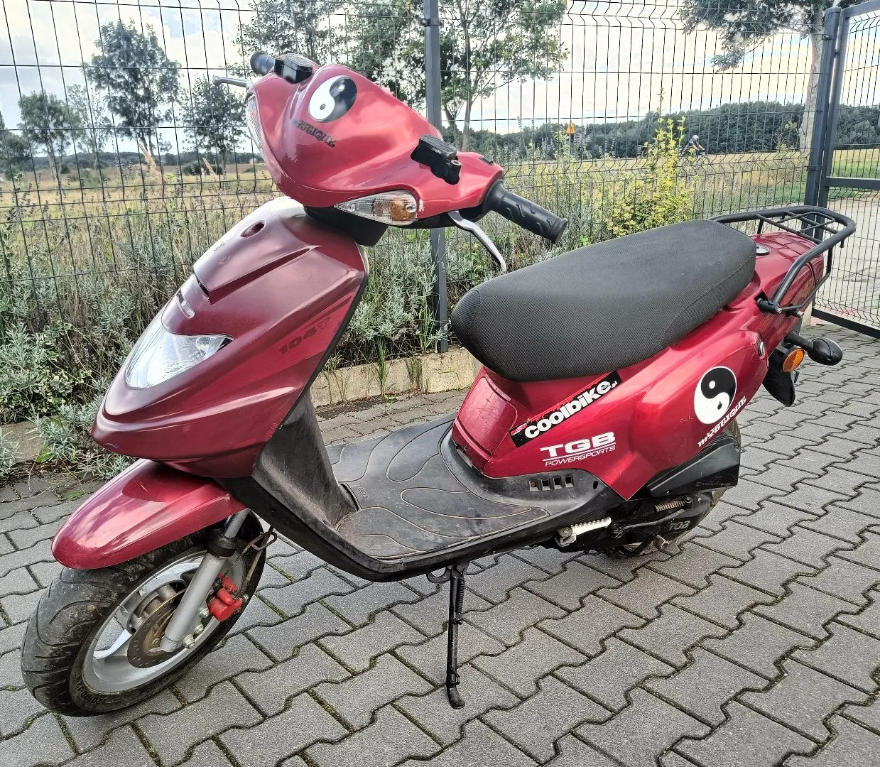 Czerwony skuter marki TGB zaparkowany na kostce brukowej przed metalowym ogrodzeniem. Na przedniej owiewce oraz na boku skutera znajduje się symbol yin-yang. Na boku widnieją napisy: 'coolbike.', 'TGB' oraz 'POWERSPORTS'. Skuter stoi na stopce bocznej.