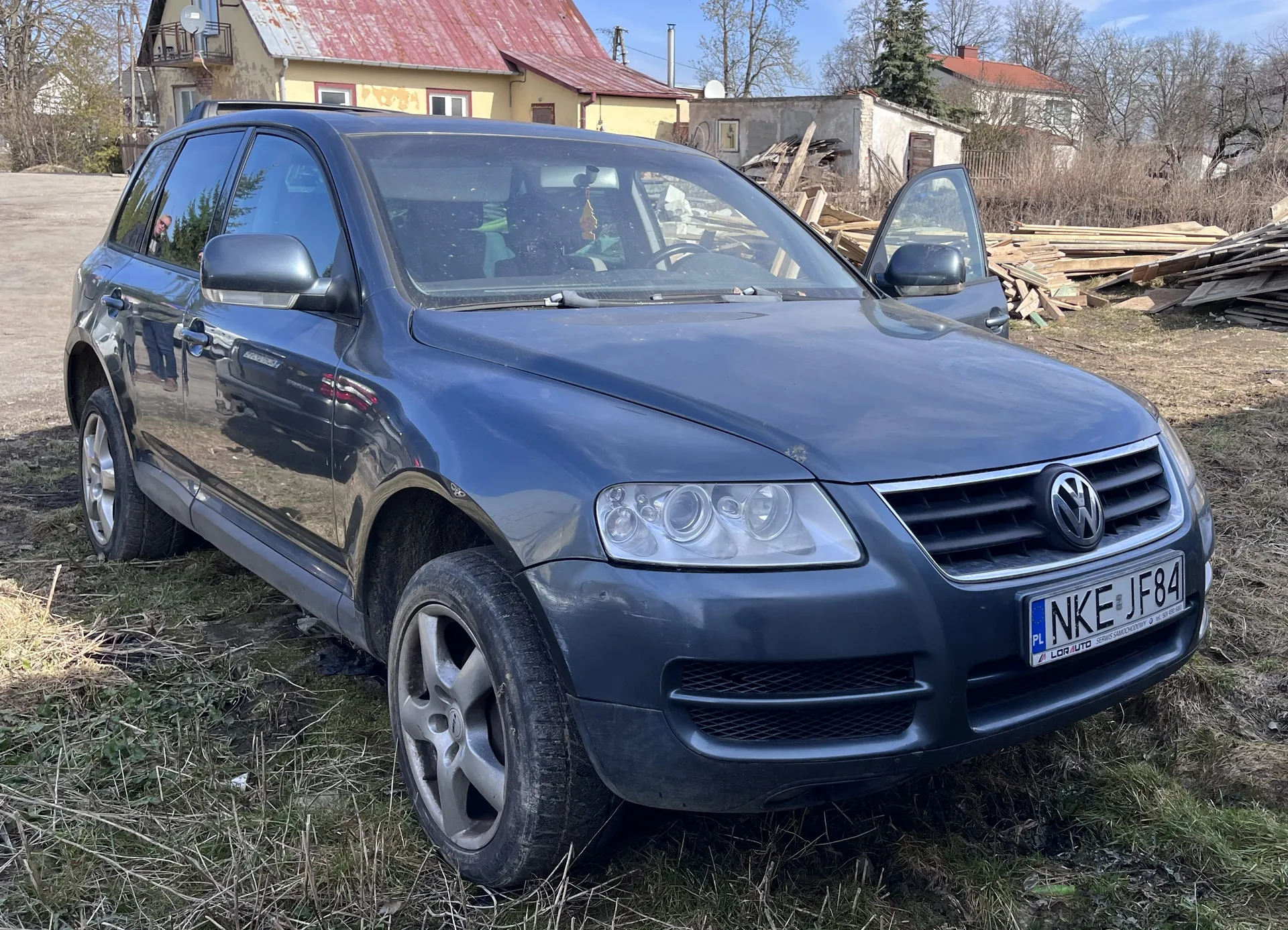 Ciemnoniebieski samochód osobowy marki Volkswagen typu SUV zaparkowany na trawiastym terenie. Przednia lewa drzwi są otwarte. Widoczna tablica rejestracyjna z polskim kodem PL i numerem NKE JF84 oraz napisem LOR AUTO na ramce. W tle domy z czerwonymi dachami, sterta desek i suche krzewy. Niebo jest jasne, pogoda słoneczna.