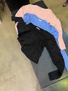 Trzy swetry w kolorach różowym, niebieskim i czarnym ułożone na ciemnej powierzchni, na tle podłogi z kafelkami.