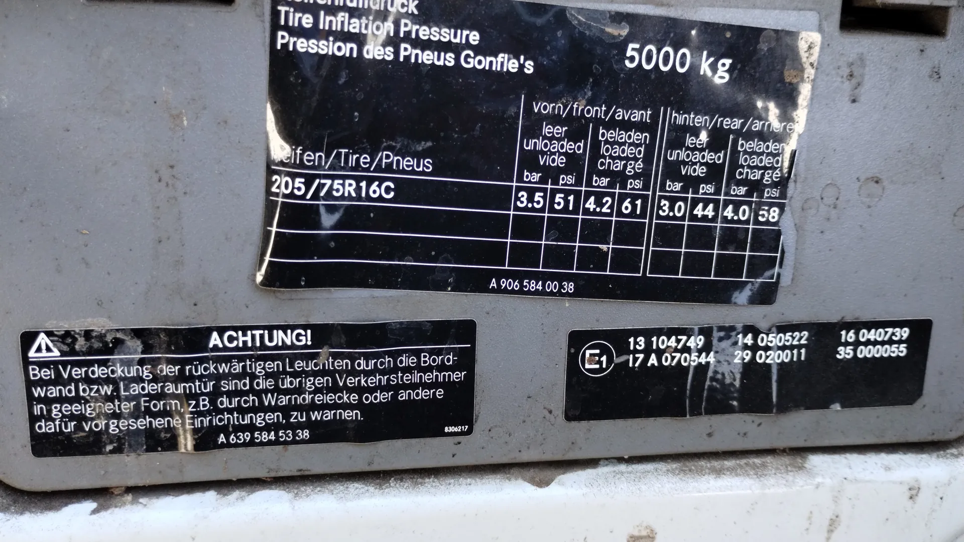 Tabliczka z informacjami o ciśnieniu w oponach. Na czarnym tle białym tekstem: 'Tire Inflation Pressure', 'Pression des Pneus Gonfle's', '5000 kg'. Wskazania dla przodu (vorn/front/avant) i tyłu (hinten/rear/arriere), wersje 'leer/unloaded/vide' i 'beladen/loaded/chargé' z wartościami w bar i psi: przód 3.5 bar/51 psi (pusty), 4.2 bar/61 psi (załadowany); tył 3.0 bar/44 psi (pusty), 4.0 bar/58 psi (załadowany). Rozmiar opony: 205/75R16C. Poniżej tabliczka ostrzegawcza z napisem 'ACHTUNG!' i niemieckim tekstem: 'Bei Verdeckung der rückwärtigen Leuchten durch die Bordwand bzw. Laderaumtür sind die übrigen Verkehrsteilnehmer in geeigneter Form, z.B. durch Warndreiecke oder andere dafür vorgesehene Einrichtungen, zu warnen.' Kod: A 639 584 53 38. Po prawej tabliczka z oznaczeniem 'E1' i numerami: 13 104749, 14 050522, 16 040739, 17 A 070544, 29 020011, 35 000055. Na głównej tabliczce kod: A 906 584 00 38. Tło: metalowa powierzchnia z widocznymi śladami zużycia.