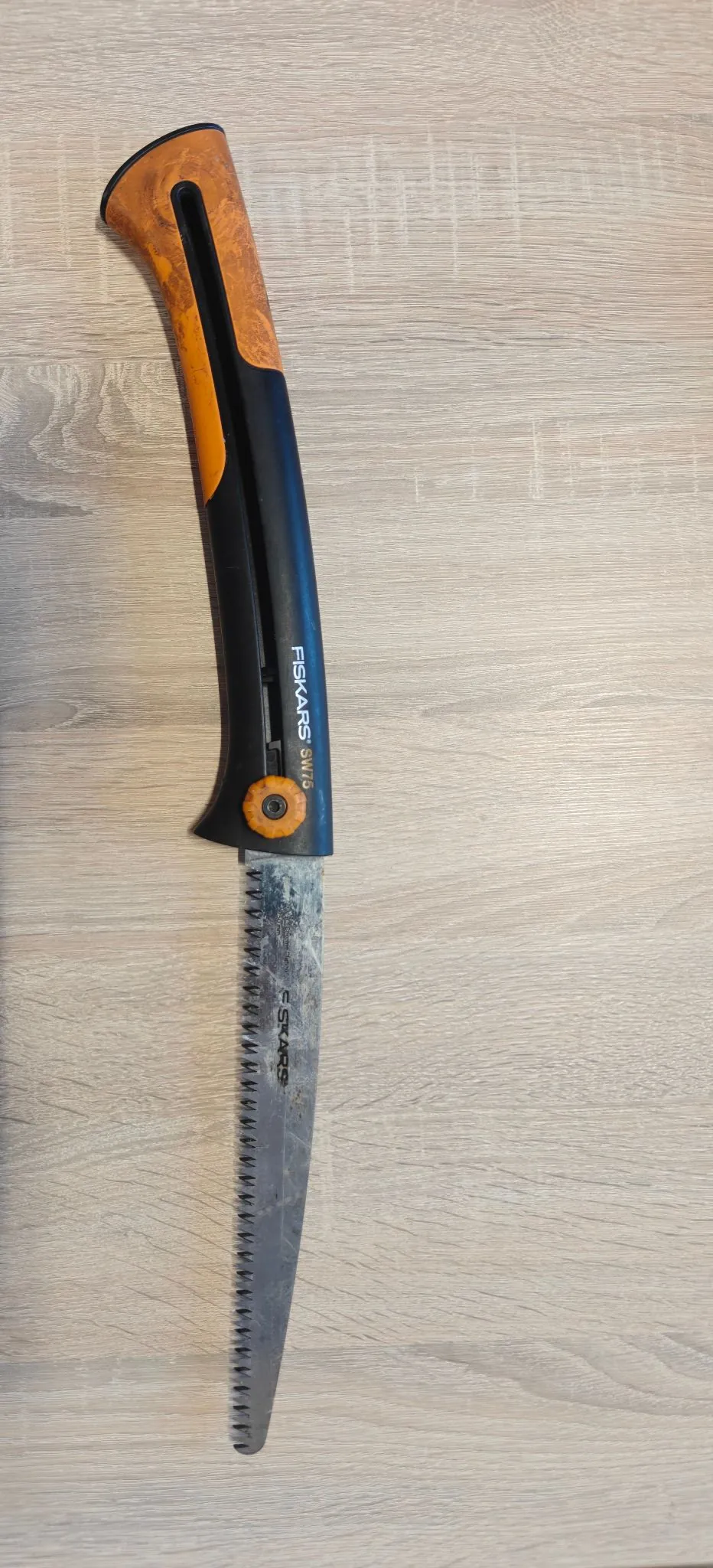 Piła ręczna marki Fiskars model SW70 leżąca pionowo na jasnym, drewnianym blacie. Narzędzie posiada czarno-pomarańczową rękojeść z napisem 'FISKARS SW70' oraz długie, wąskie, ząbkowane ostrze wykonane ze stali.