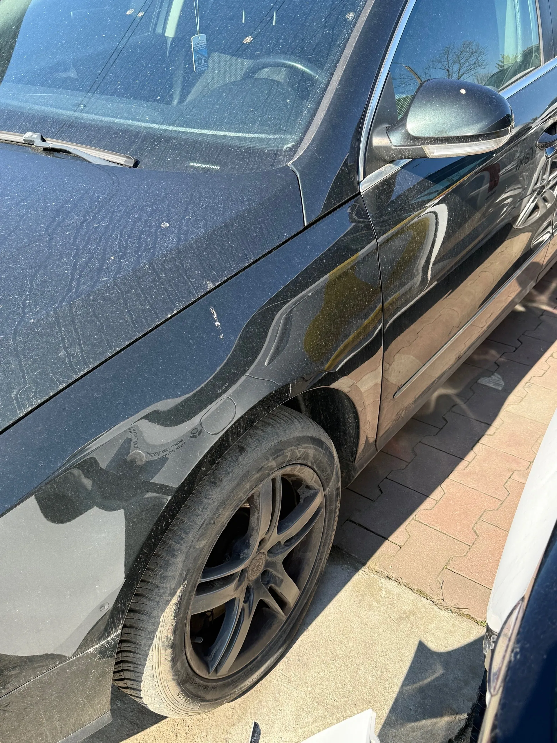 Czarny samochód widziany z boku i przodu, z widocznym przednim kołem, lusterkiem bocznym oraz przednią szybą. Na przedniej szybie wiszący jest mały znaczek. Samochód stoi na brukowanej powierzchni, a na jego karoserii widoczne są ślady kurzu i drobnych zadrapań. W tle częściowo widoczny jest inny pojazd oraz drzewa.