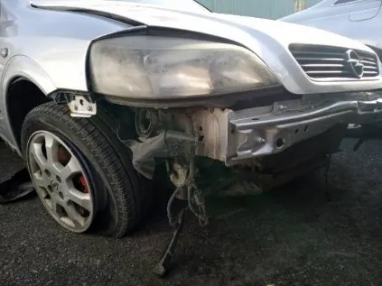 Zdjęcie przedstawia uszkodzony przód srebrnego samochodu. Widoczne są zniszczone elementy: brakujący zderzak, pęknięte reflektory, odsłonięte części podwozia oraz koło z aluminiową felgą. W tle częściowo widoczny jest inny pojazd. Samochód stoi na asfaltowej powierzchni.