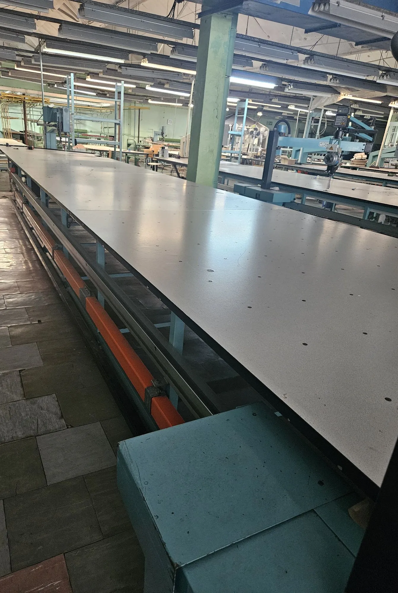 Wnętrze hali przemysłowej z długim, metalowym stołem montażowym lub linią produkcyjną o szarej powierzchni. Pod blatem widoczna jest konstrukcja wsporcza z niebieskimi profilami i pomarańczowymi elementami. W tle znajdują się inne maszyny warsztatowe, niebieskie słupy konstrukcyjne oraz oświetlenie liniowe zamontowane pod sufitem.