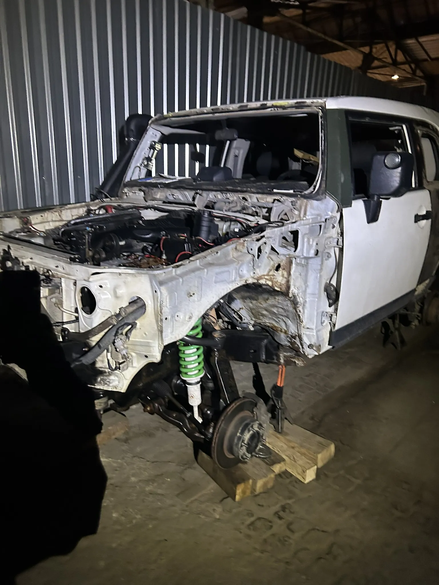 Zdjęcie przedstawia uszkodzony samochód terenowy w warsztacie. Pojazd ma białą karoserię z zielonymi elementami, brakuje mu maski i części przedniego nadwozia, co odsłania silnik oraz podzespoły. Widoczna jest zielona sprężyna zawieszenia, a przednia oś spoczywa na drewnianych podkładkach. W tle metalowa ściana z pionowymi listwami i betonowa podłoga. Samochód znajduje się wewnątrz hali z metalowym dachem.