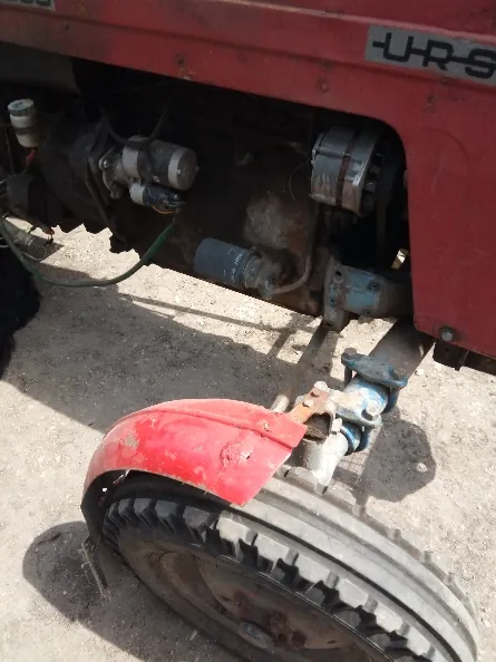 Zbliżenie na fragment czerwonego traktora, przedstawiające silnik, układ elektryczny z alternatorem oraz przednie koło z czerwonym błotnikiem. Na obudowie traktora widoczny jest fragment napisu „-URS”.