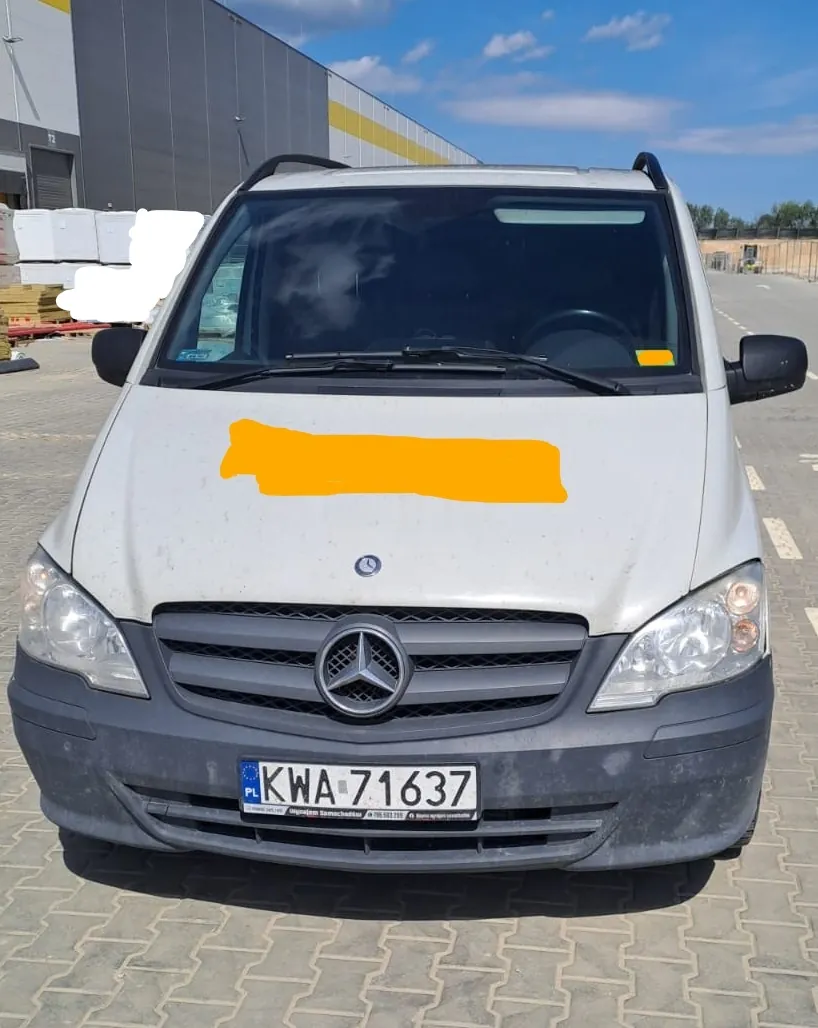 Srebrny samochód dostawczy marki Mercedes-Benz zaparkowany na kostce brukowej przed budynkiem magazynowym. Na masce pojazdu znajduje się pomarańczowy zakryty obszar. Samochód ma polską tablicę rejestracyjną o numerze KWA 71637.