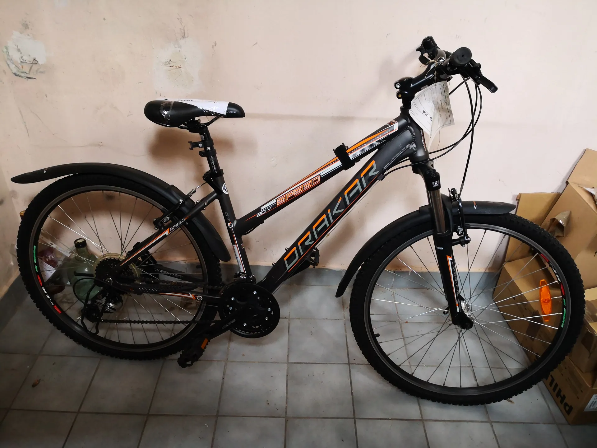 Czarny rower górski marki DRAKAR z pomarańczowymi i białymi elementami, model SPEED. Rower stoi na szarych płytkach podłogowych, oparty o beżową ścianę z widocznymi uszkodzeniami. Na ramie widoczny napis DRAKAR oraz SPEED. Przednie koło ma pomarańczowy reflektor, a na kierownicy zawieszony jest biały dokument. Po prawej stronie obrazu znajdują się brązowe kartony. Siedzisko jest czarne z białą etykietą.