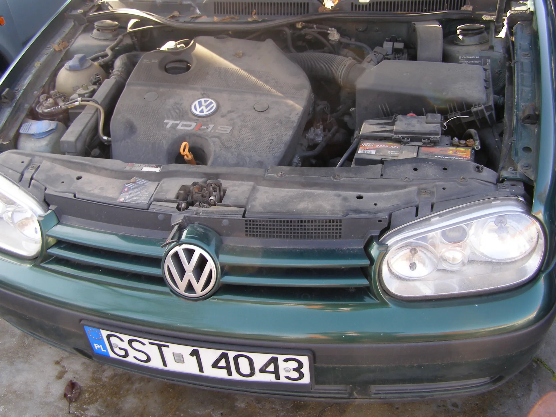 Zielony samochód marki Volkswagen z otwartą maską silnika. Widoczny silnik z napisem TDI 1.9 oraz logo VW na pokrywie silnika. Na przednim zderzaku umieszczona tablica rejestracyjna z polskim znakiem PL i numerem GST 14043. Widoczne są również reflektory, logo VW na atrapie chłodnicy oraz elementy silnika, w tym akumulator i przewody.