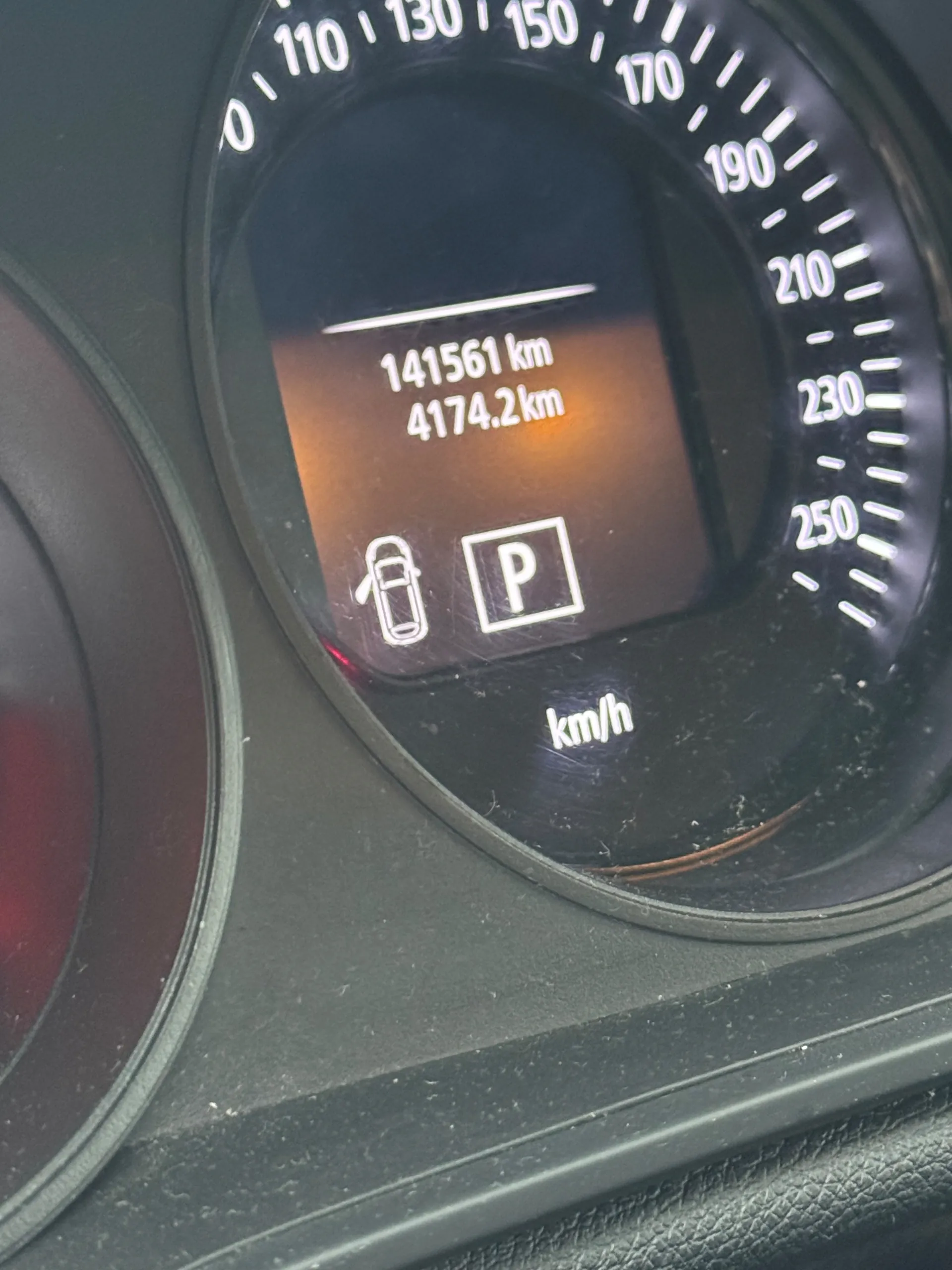 Zbliżenie na samochodowy licznik prędkości z czarnym tłem. Na wyświetlaczu cyfrowym widoczne są wartości: 141561 km oraz 4174.2 km. Poniżej znajduje się ikona samochodu z pasem bezpieczeństwa i kwadrat z literą P. Skala prędkości w km/h z białymi cyframi od 0 do 250, oznaczenie jednostki km/h na dole. Licznik pokryty jest lekkim kurzem.
