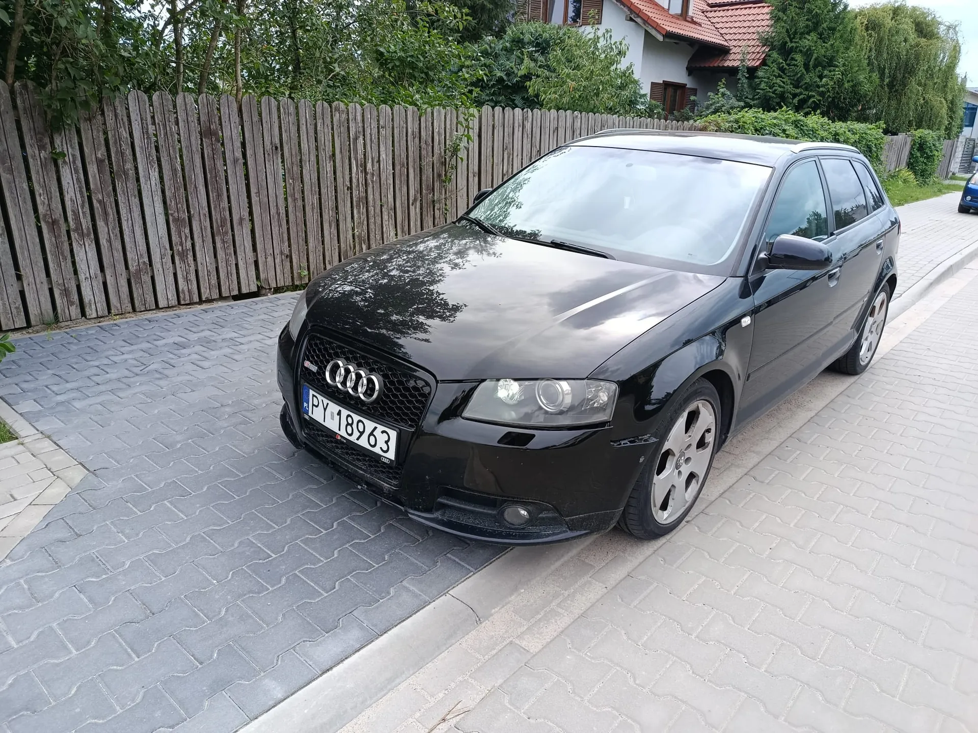 Czarny samochód marki Audi A3 zaparkowany na szarym, kostkowanym podjeździe obok drewnianego ogrodzenia. Samochód jest ustawiony przodem do widza, widoczna jest tablica rejestracyjna o numerze PY 18963. W tle widać zielone drzewa oraz fragment domu z czerwonym dachem.