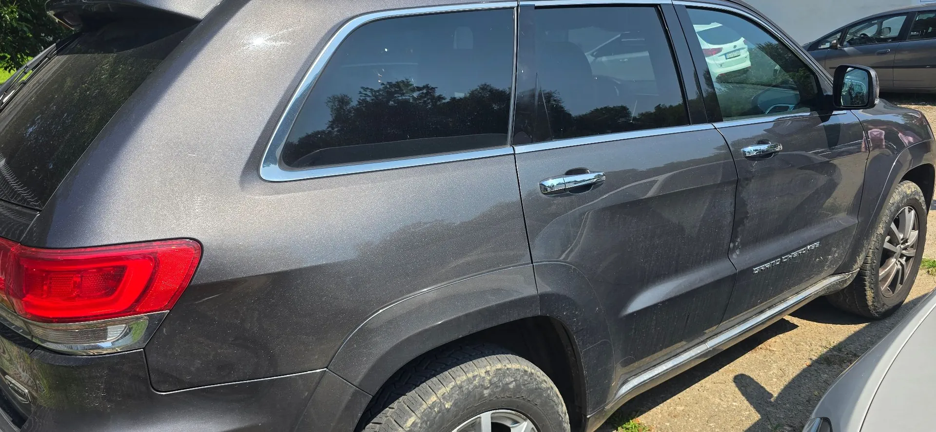Boczny widok tylnej części ciemnoszarego samochodu typu SUV. Widoczna jest tylna lampa, tylne i boczne szyby z chromowanymi obramowaniami oraz klamki. Na drzwiach widnieje napis 'GRAND CHEROKEE'. Samochód jest lekko zakurzony, szczególnie w dolnych partiach nadkoli.