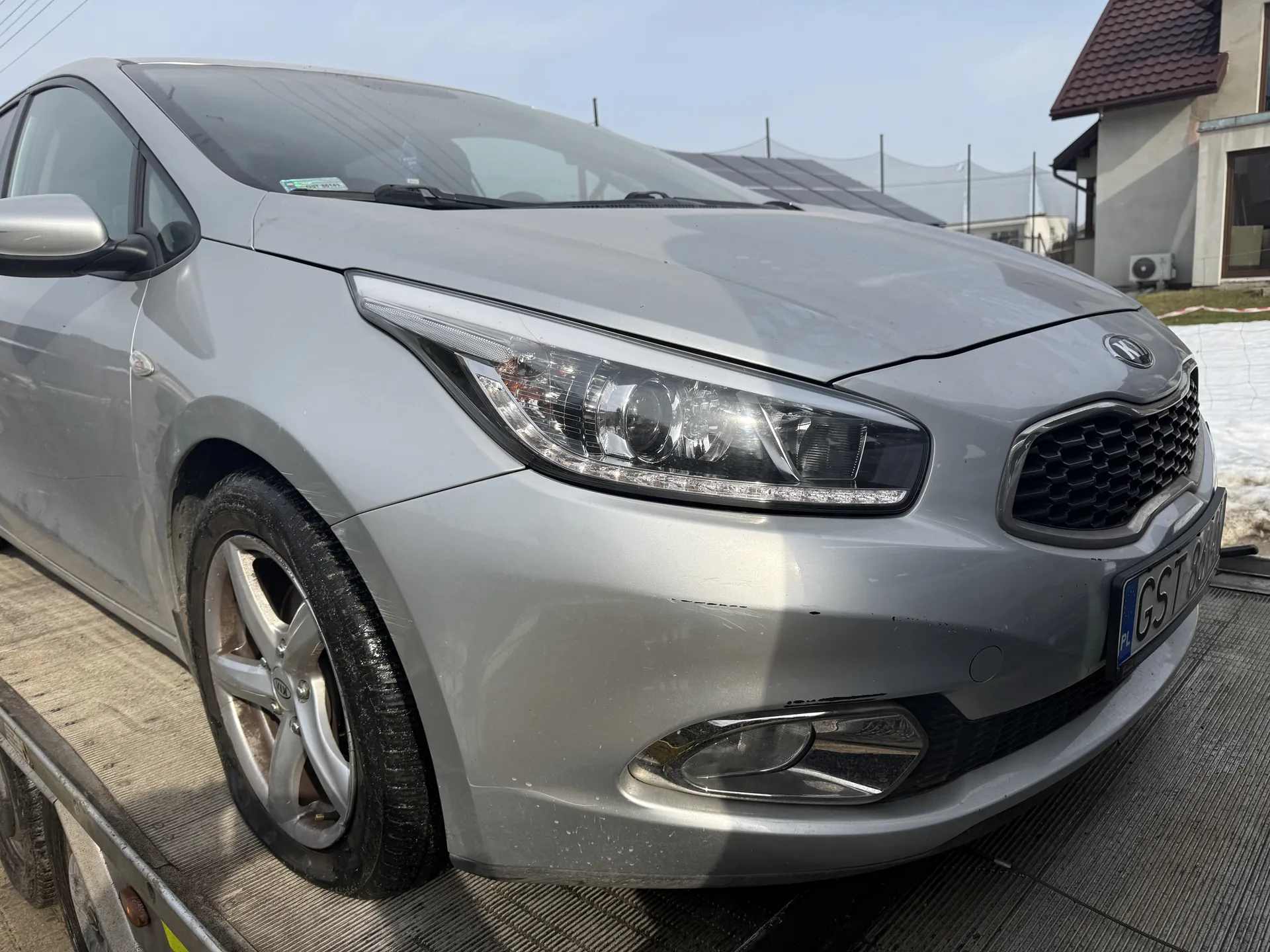 Srebrny samochód marki Kia z przodu, stojący na metalowej platformie przyczepy. Widoczne są reflektory, atrapa chłodnicy, tablica rejestracyjna z napisem GST oraz fragmentem numeru. W tle dom z czerwoną dachówką, panele słoneczne i resztki śniegu na ziemi. Na przedniej szybie samochodu znajduje się mały naklejony dokument.