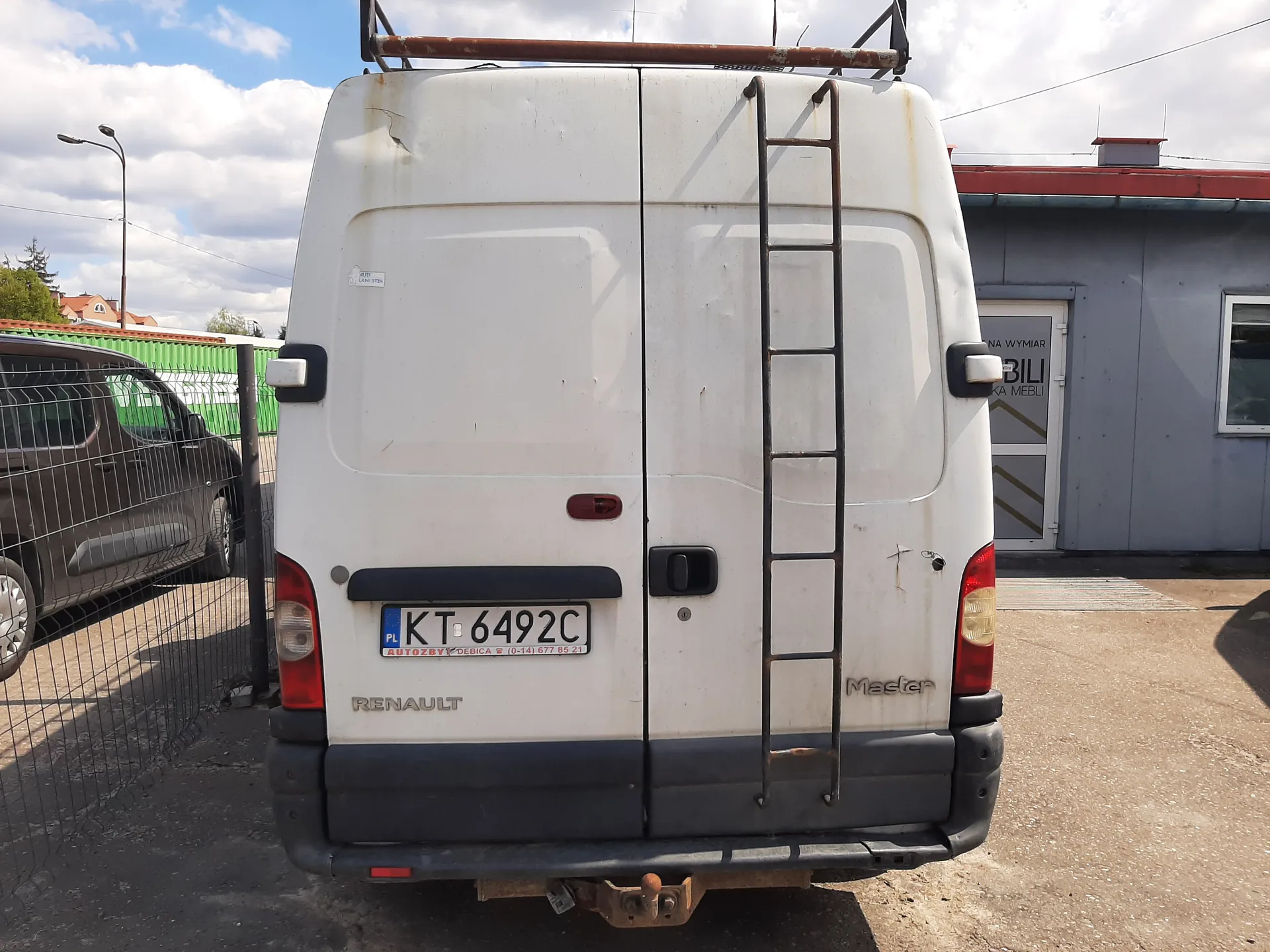 Biały samochód dostawczy Renault Master widziany od tyłu. Na tylnych drzwiach umocowana jest metalowa drabina. Widoczny jest polski numer rejestracyjny KT 6492C z napisem "AUTOZBYT DEBICA" i numerem telefonu poniżej. Po lewej stronie obrazu znajduje się czarna siatka ogrodzeniowa oraz brązowy samochód osobowy. W tle budynek z napisem "NA WYMIAR BILI KA MEBLI" na drzwiach, zielona bariera i niebo z chmurami. Samochód stoi na asfaltowej powierzchni.
