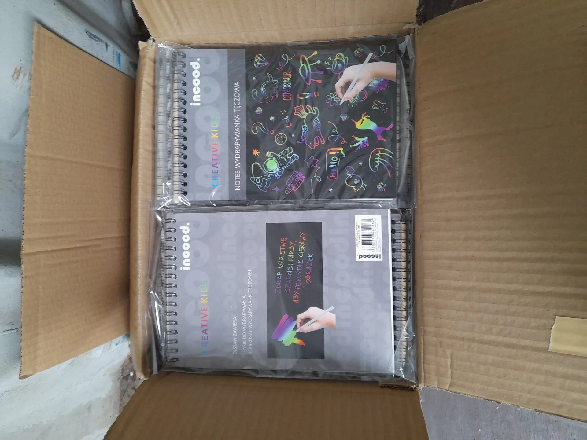 Dwa spiralne zeszyty marki 'incood.' z serii 'CREATIVE KIDS' leżące w kartonowym pudełku, zapakowane w przezroczystą folię. Górny zeszyt ma okładkę z kolorowymi rysunkami na czarnym tle i napisem 'NOTATNIK WYDRAPYWANKA TĘCZOWA'. Dolny zeszyt zawiera opis funkcji produktu, w tym informację o zawartości: '1 RYŁCEK DO WYGRAPYWANIA' oraz '10 ARKUSZY WYDRAPYWANKI TĘCZOWEJ'. Na okładkach widoczne są grafiki przedstawiające dłoń z rylcem wydrapującym kolorowy wzór.