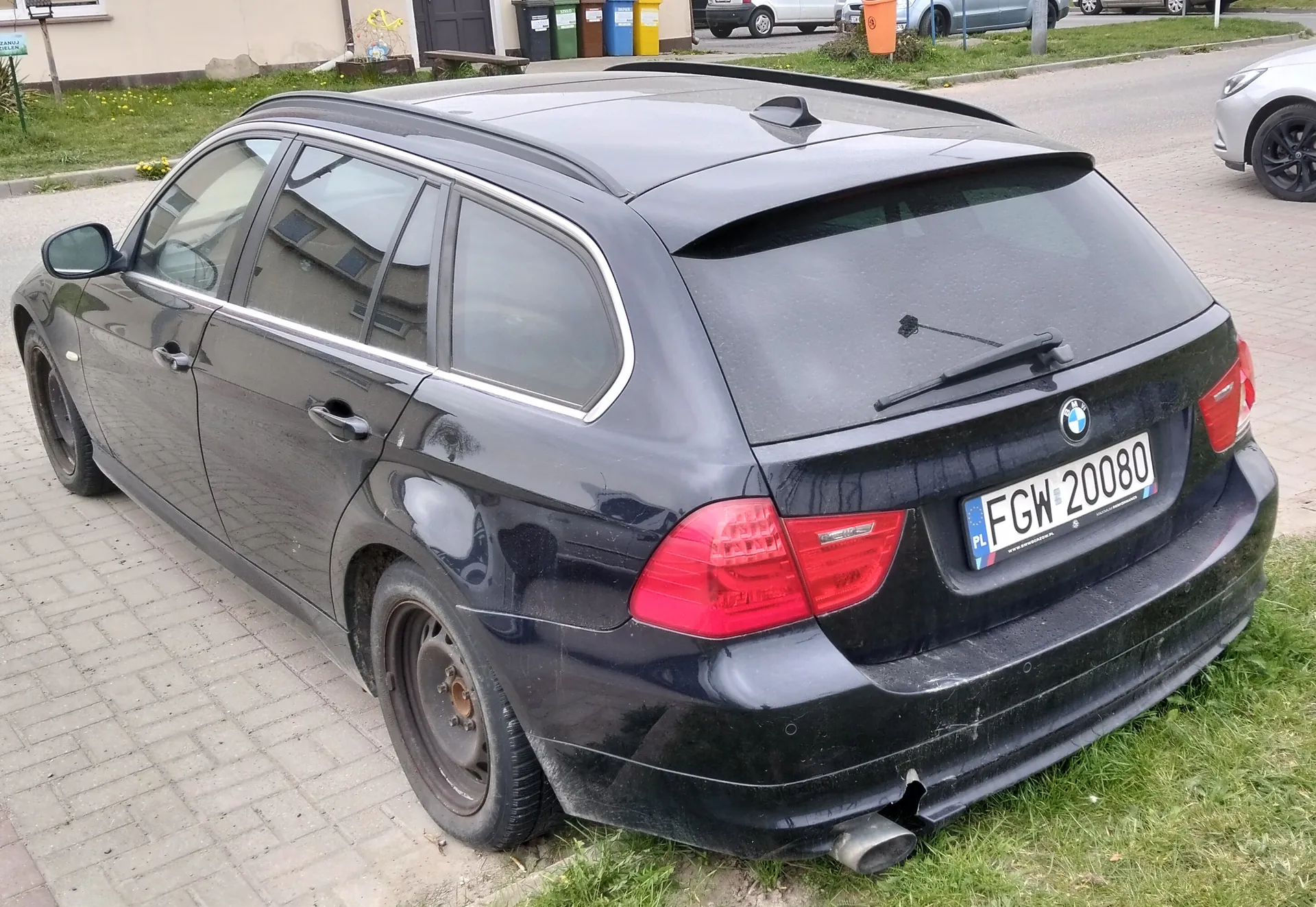 Czarny samochód osobowy typu kombi marki BMW zaparkowany na brukowanej powierzchni. Widok od tyłu z lewej strony. Na tablicy rejestracyjnej widoczny napis PL FGW 20080. Samochód posiada dachowy bagażnik, czerwone światła tylne oraz uszkodzony tylny zderzak. W tle budynki, kolorowe pojemniki na śmieci, trawnik i inne zaparkowane pojazdy.