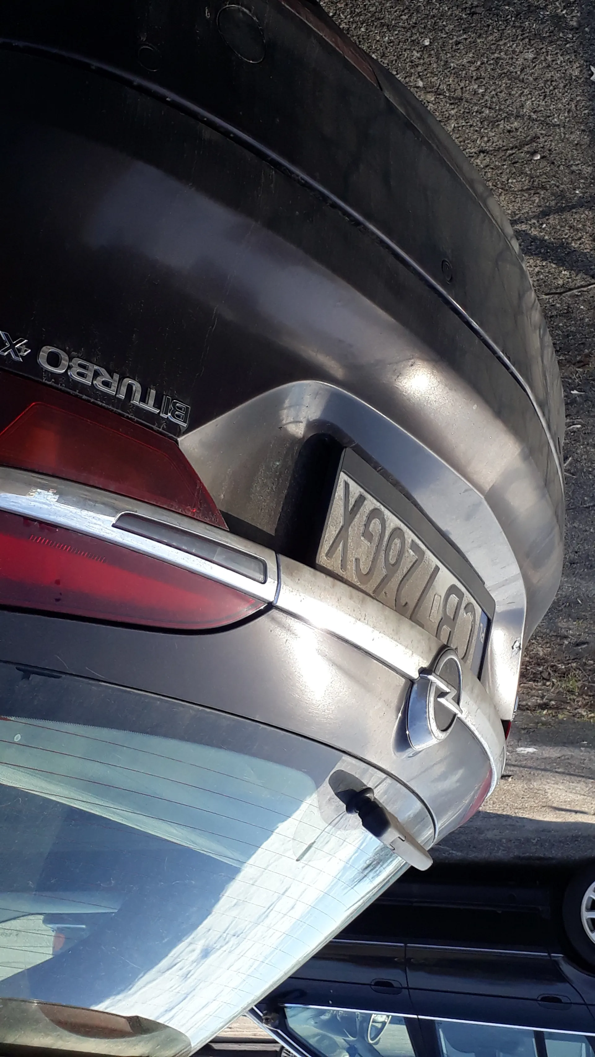 Zbliżenie na tylną część ciemnoszarego samochodu z widocznym czerwonym światłem tylnym, napisem 'BITURBO 4X' oraz tablicą rejestracyjną 'CB 126GX'. Po lewej stronie częściowo widoczny inny czarny samochód. Tło stanowi asfaltowa powierzchnia.