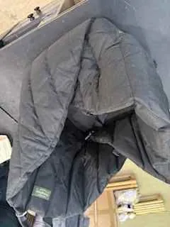 Ciemnoszara pikowana kurtka leżąca na powierzchni. W tle widoczne są drewniane elementy konstrukcyjne oraz częściowo widoczne przedmioty, w tym biały obiekt i metalowe elementy po prawej stronie. Na kurtce znajduje się zielona etykieta z nieczytelnym tekstem.