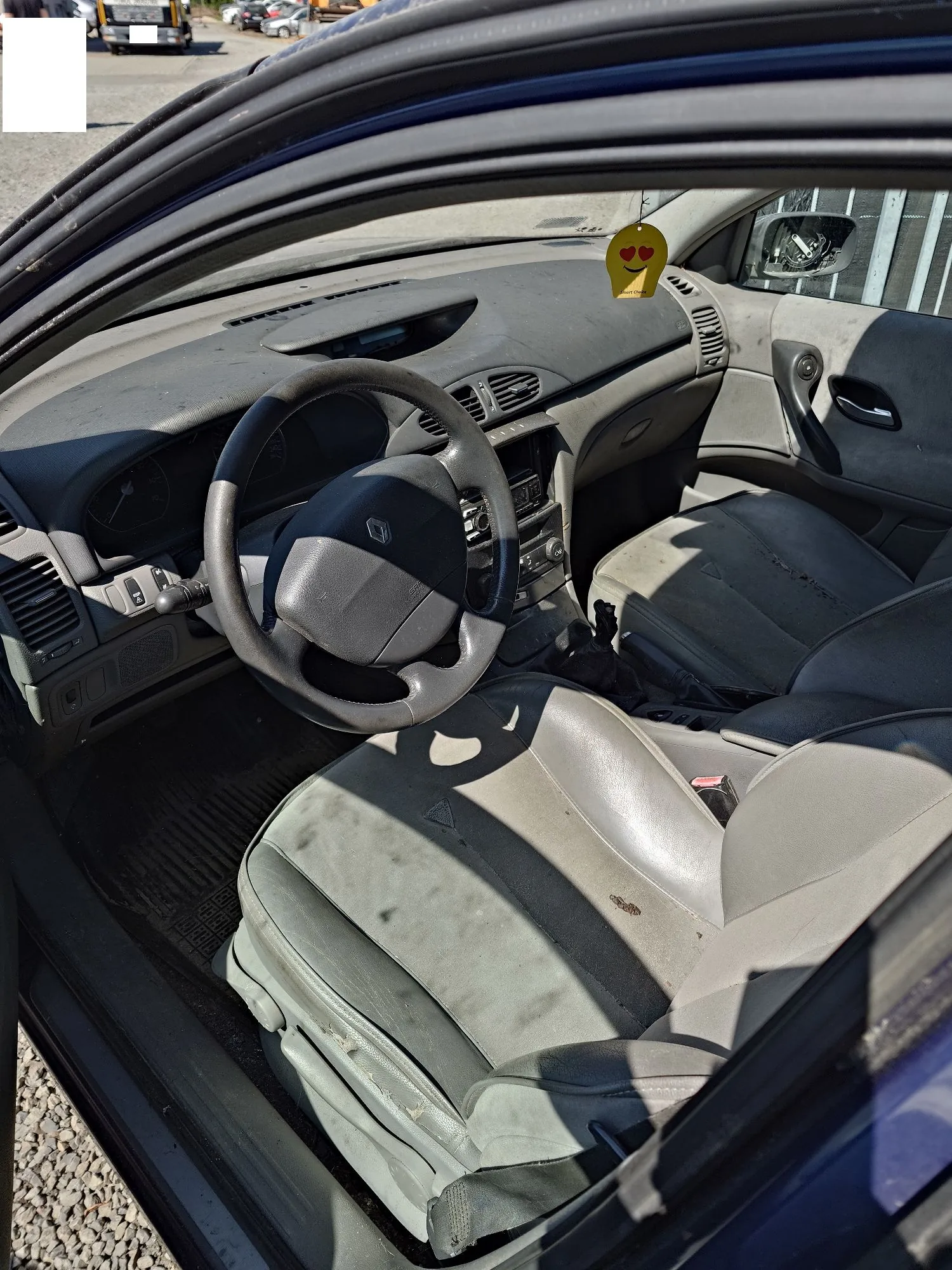 Wnętrze samochodu z kierownicą po prawej stronie. Widoczna jest szara deska rozdzielcza, kierownica z logo marki Renault, dwa przednie fotele z szarej tkaniny oraz fragment drzwi pasażera. Na desce rozdzielczej znajduje się żółty, okrągły zawieszkowy gadżet z emotikoną zakochanej twarzy. Wnętrze jest zakurzone.