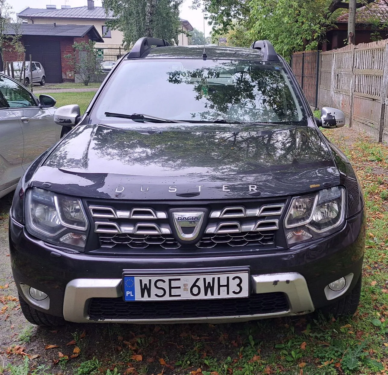 Czarny samochód marki Dacia Duster zaparkowany na trawiastym podjeździe przed domem. Widoczny jest przód pojazdu z tablicą rejestracyjną o numerze WSE 6WH3. Na masce znajduje się napis DUSTER, a na grillu logo DACIA. Samochód posiada relingi dachowe.