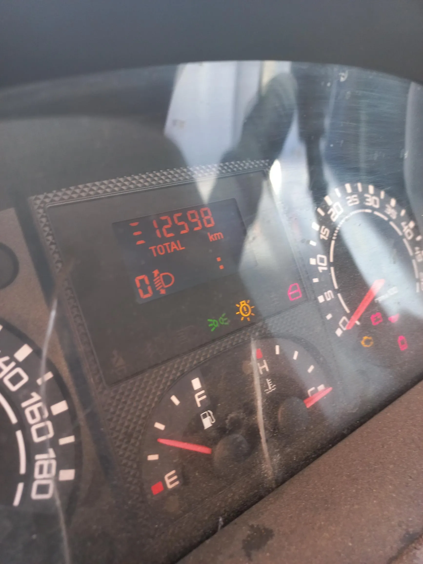 Zbliżenie na deskę rozdzielczą samochodu. W centralnej części znajduje się cyfrowy wyświetlacz z pomarańczowym tekstem: '12598 km TOTAL' oraz ikoną lampy i cyfrą '0'. Poniżej wyświetlacza świecą się żółte kontrolki świateł. Po prawej stronie widoczny jest fragment obrotomierza z podziałką od 0 do 40, a na dole wskaźnik poziomu paliwa z oznaczeniami 'F' (full) i 'E' (empty). Po lewej stronie widoczny jest fragment prędkościomierza z wartościami 160 i 180.