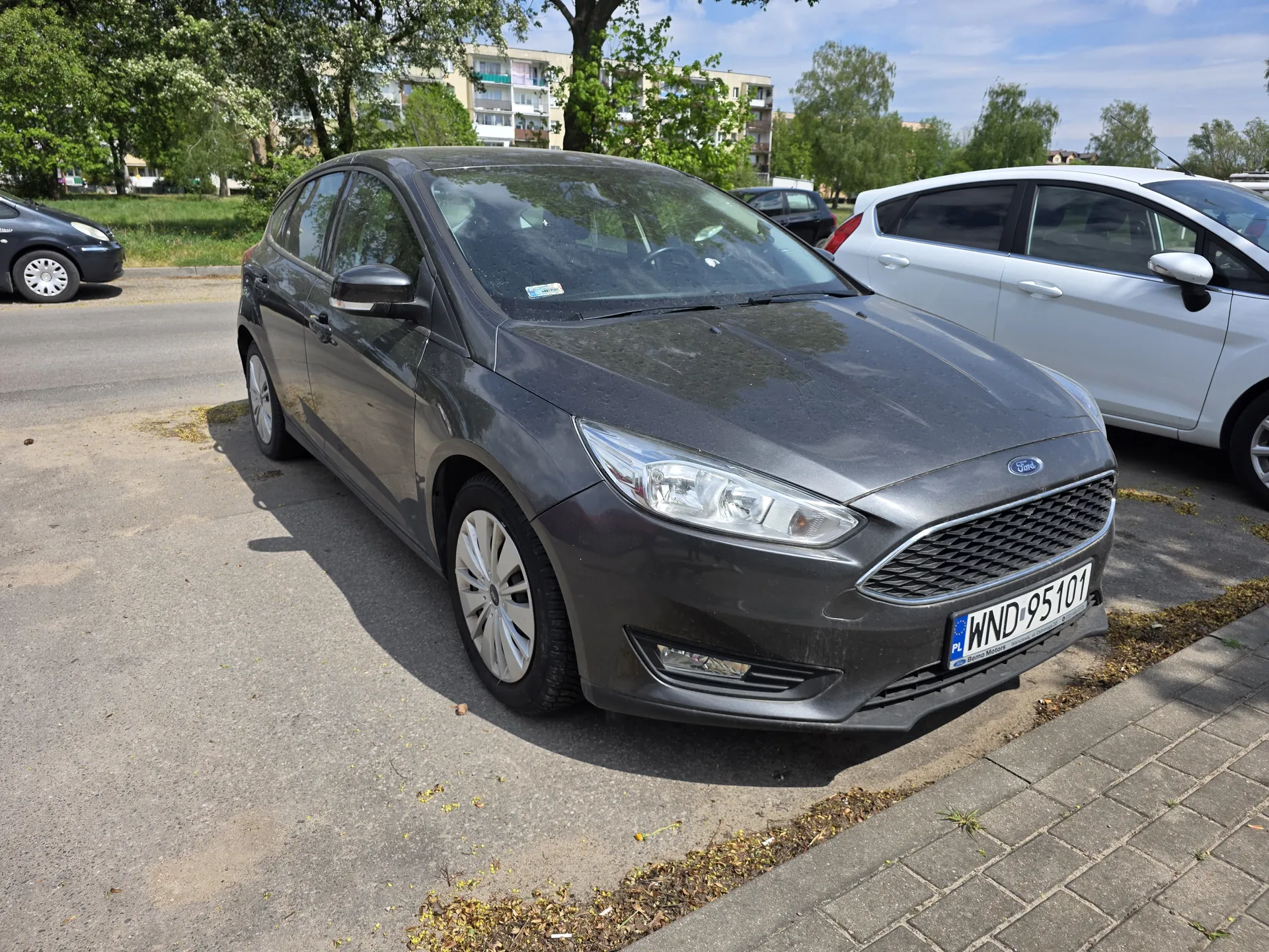 Szary samochód marki Ford z tablicą rejestracyjną WND 95101 zaparkowany na asfaltowym placu parkingowym. W tle widoczne są biały i czarny samochód, drzewa oraz bloki mieszkalne. Jasne niebo z kilkoma chmurami.