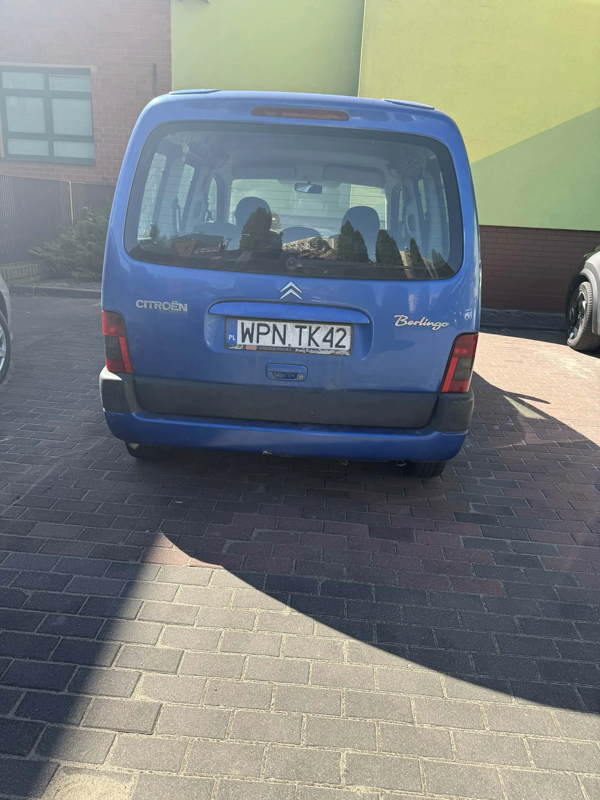 Z tyłu niebieski samochód Citroën Berlingo z tablicą rejestracyjną WPN TK42 (PL). Samochód stoi na brukowanej powierzchni. W tle widoczne są budynki z zielonymi i ceglanymi ścianami oraz fragment okna z roletami. Po prawej stronie częściowo widoczny inny pojazd. Na tylnym szybie odbijają się drzewa i budynki.