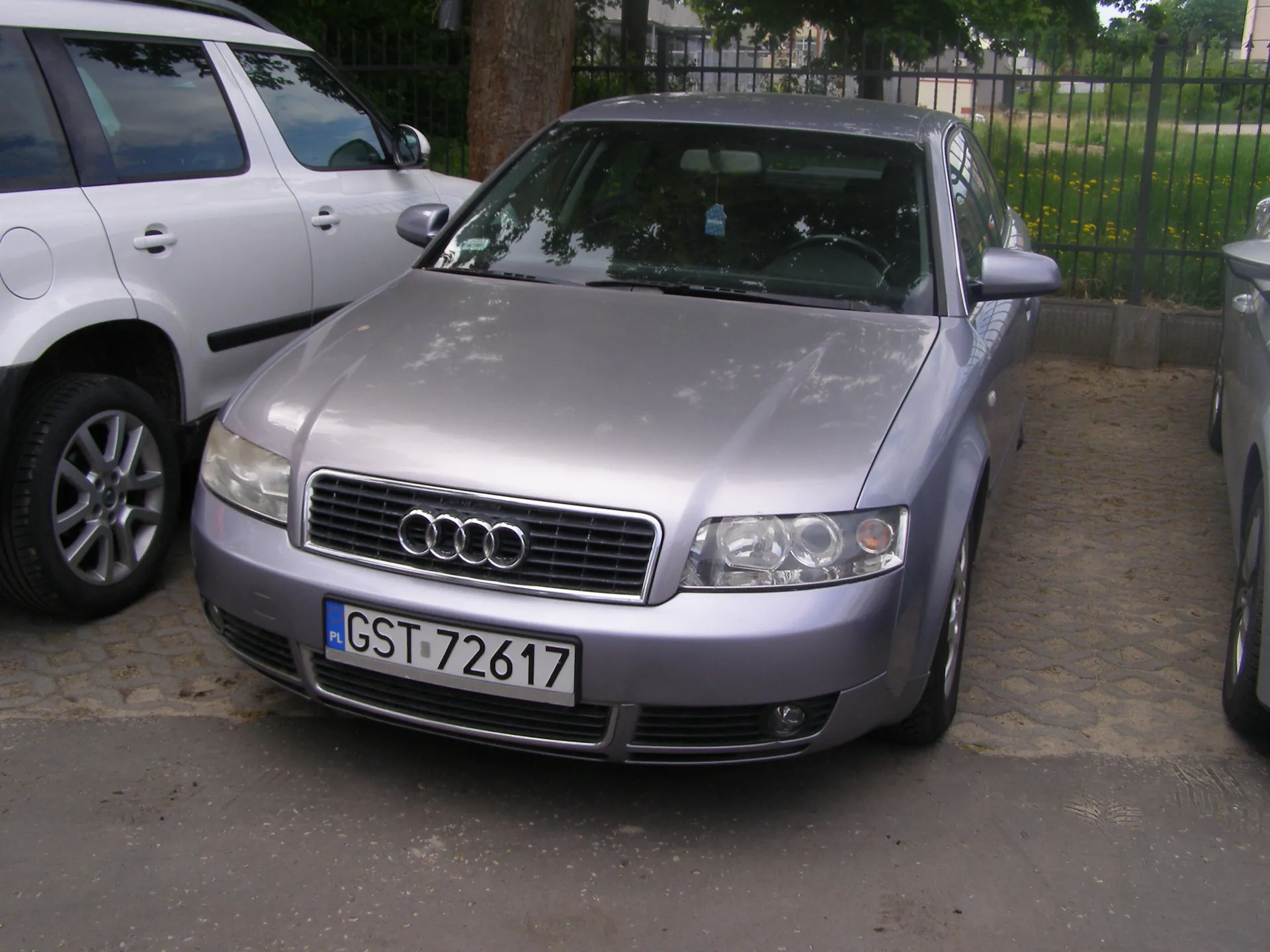 Srebrny samochód marki Audi zaparkowany na brukowanym placu. Widoczna tablica rejestracyjna z napisem GST 72617 oraz oznaczeniem PL. Po lewej stronie biały SUV, w tle czarny metalowy płot, trawa, drzewa i budynki. Samochód ma cztery pierścienie w logo na masce, reflektory i lusterka boczne.