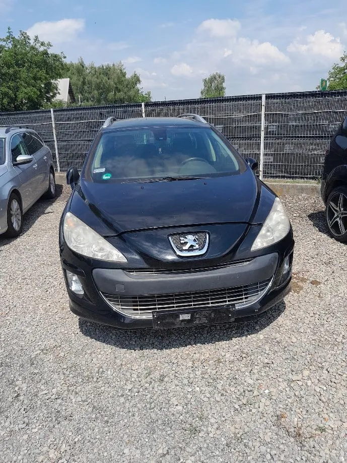 Czarny samochód marki Peugeot zaparkowany przodem do obiektywu na żwirowym placu. Po lewej stronie widoczny jest fragment srebrnego samochodu, a po prawej fragment czarnego auta. W tle znajduje się metalowy płot oraz zielone drzewa pod jasnym, lekko zachmurzonym niebem.