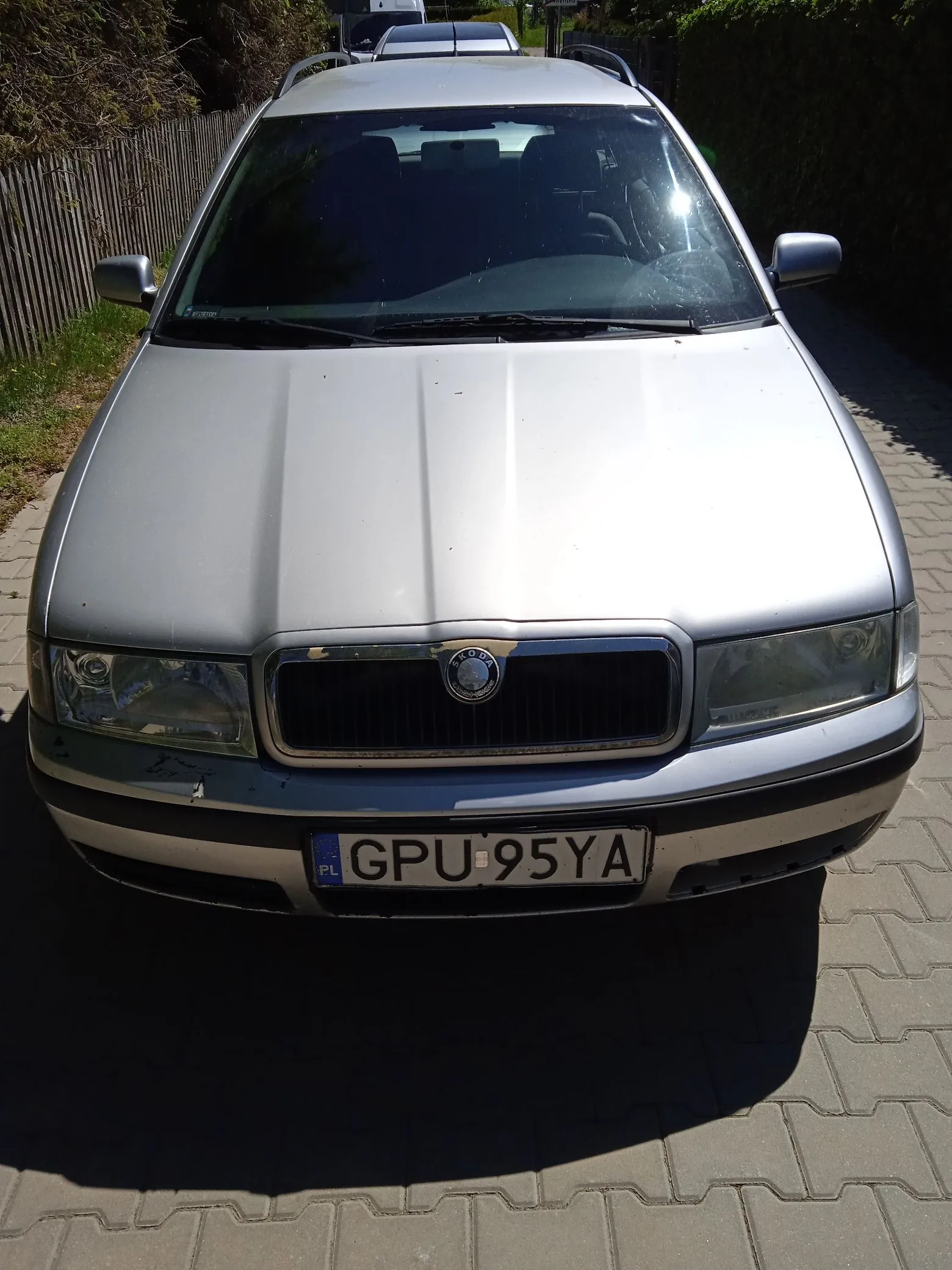 Srebrny samochód marki Skoda zaparkowany przodem do widza na kostce brukowej. Pojazd posiada bagażnik dachowy oraz polską tablicę rejestracyjną o numerze GPU 95YA. Na masce i zderzaku widoczne są drobne uszkodzenia lakieru.