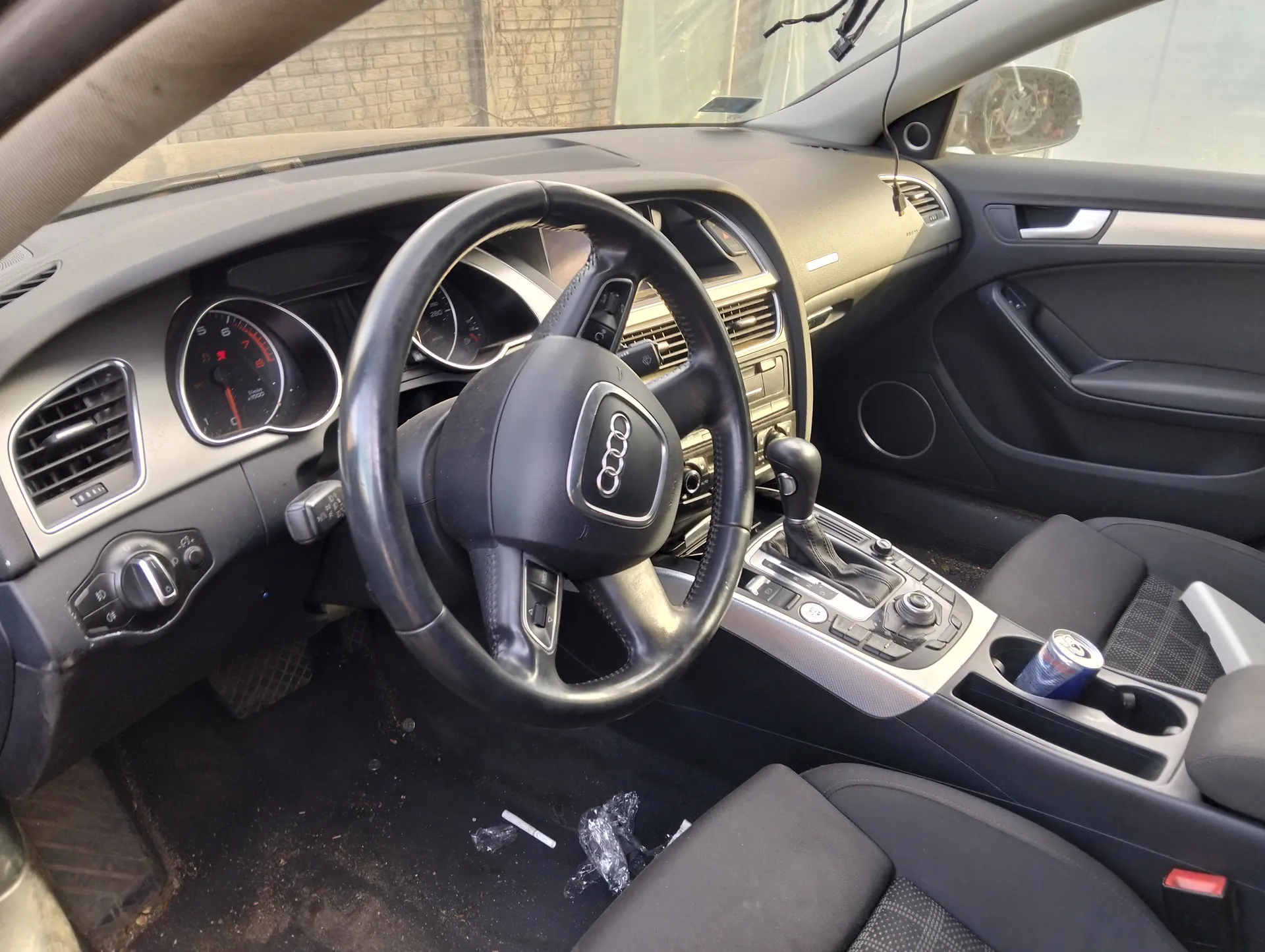 Wnętrze samochodu marki Audi z widoczną kierownicą z logo czterech pierścieni, tablicą przyrządów z licznikami, centralną konsolą z przyciskami i dźwignią zmiany biegów. W kubku na podłodze widoczny niedopałek papierosa oraz plastikowe opakowanie. W uchwycie na napoje znajduje się puszka. W tle przez przednią szybę widać ceglaną ścianę. Wnętrze posiada czarne i szare elementy wykończenia.