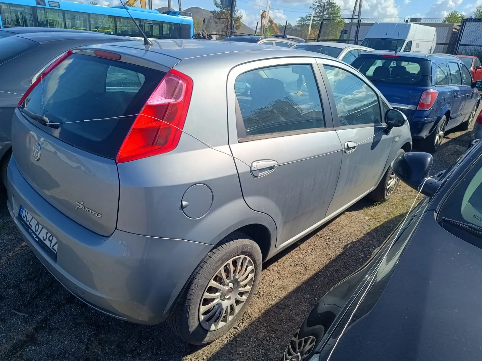 Srebrny samochód marki Fiat Punto zaparkowany na placu obok innych pojazdów. Widoczny jest tył i prawy bok auta, tablica rejestracyjna o numerze BCZ 67342 oraz napis PUNTO na klapie bagażnika. W tle widać niebieski autobus oraz białą furgonetkę.