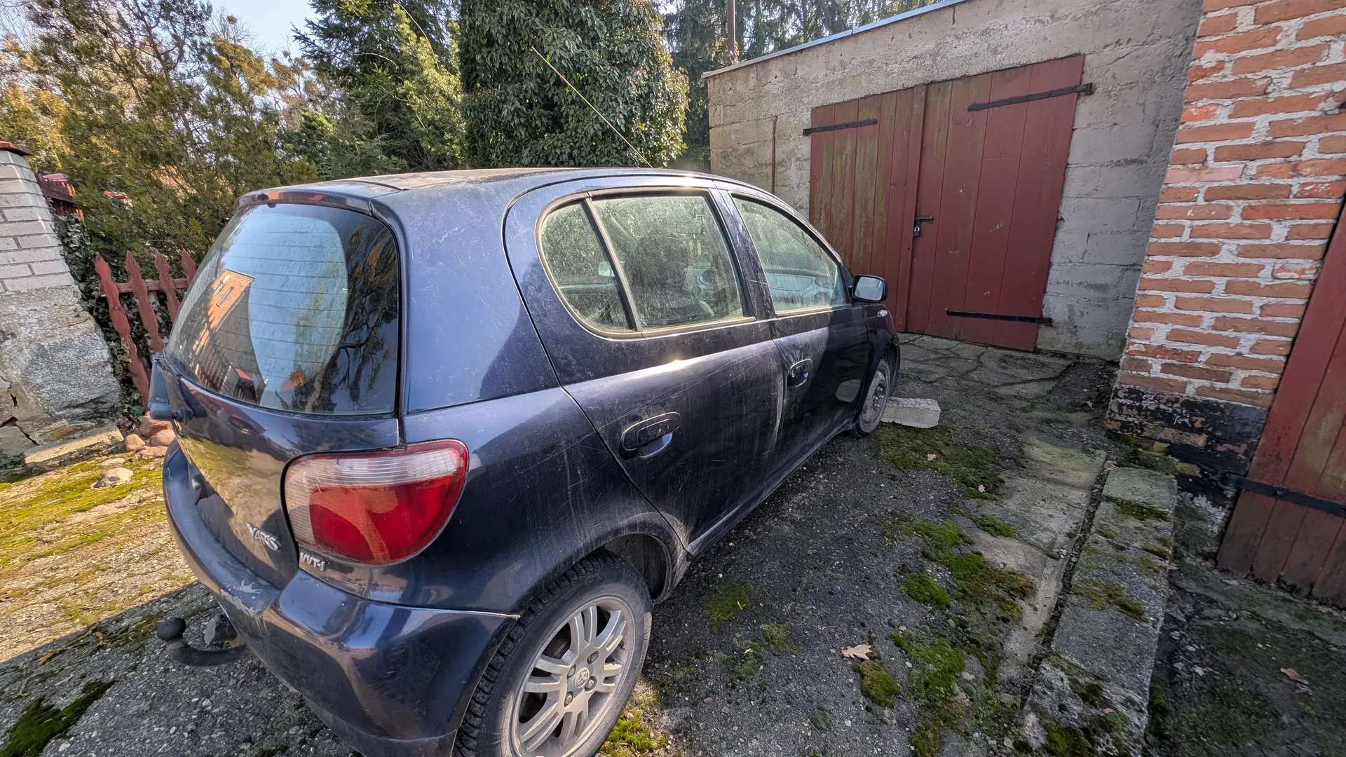 Ciemnoniebieski samochód osobowy typu hatchback (Toyota Yaris z napisem VVT-i na tylnej szybie) zaparkowany na podwórku. Samochód jest zabrudzony, z widocznymi zadrapaniami. W tle betonowa budowla z czerwonymi drzwiami garażowymi, ceglana ściana po prawej stronie, zielone drzewa i czerwony płot po lewej. Podłoga z kamieni i asfaltu porośnięta mchem. Na tylnej szybie odbicie budynku.