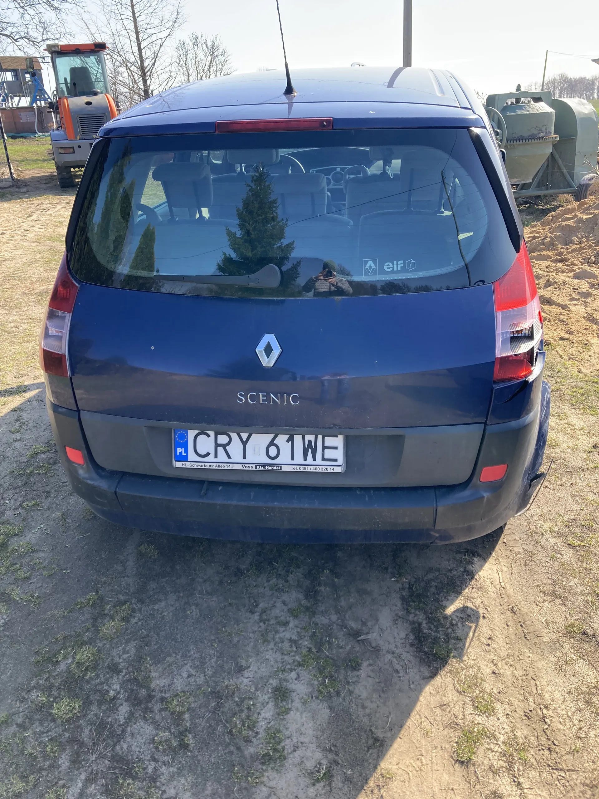 Niebieski samochód Renault Scenic widziany od tyłu. Widoczna tablica rejestracyjna z numerem CRY 61WE oraz oznaczeniem PL. Na szybie tylnej naklejka z napisem 'elf'. Samochód zaparkowany na brudnej, częściowo zarośniętej ziemi. W tle maszyna rolnicza i drzewa bez liści.