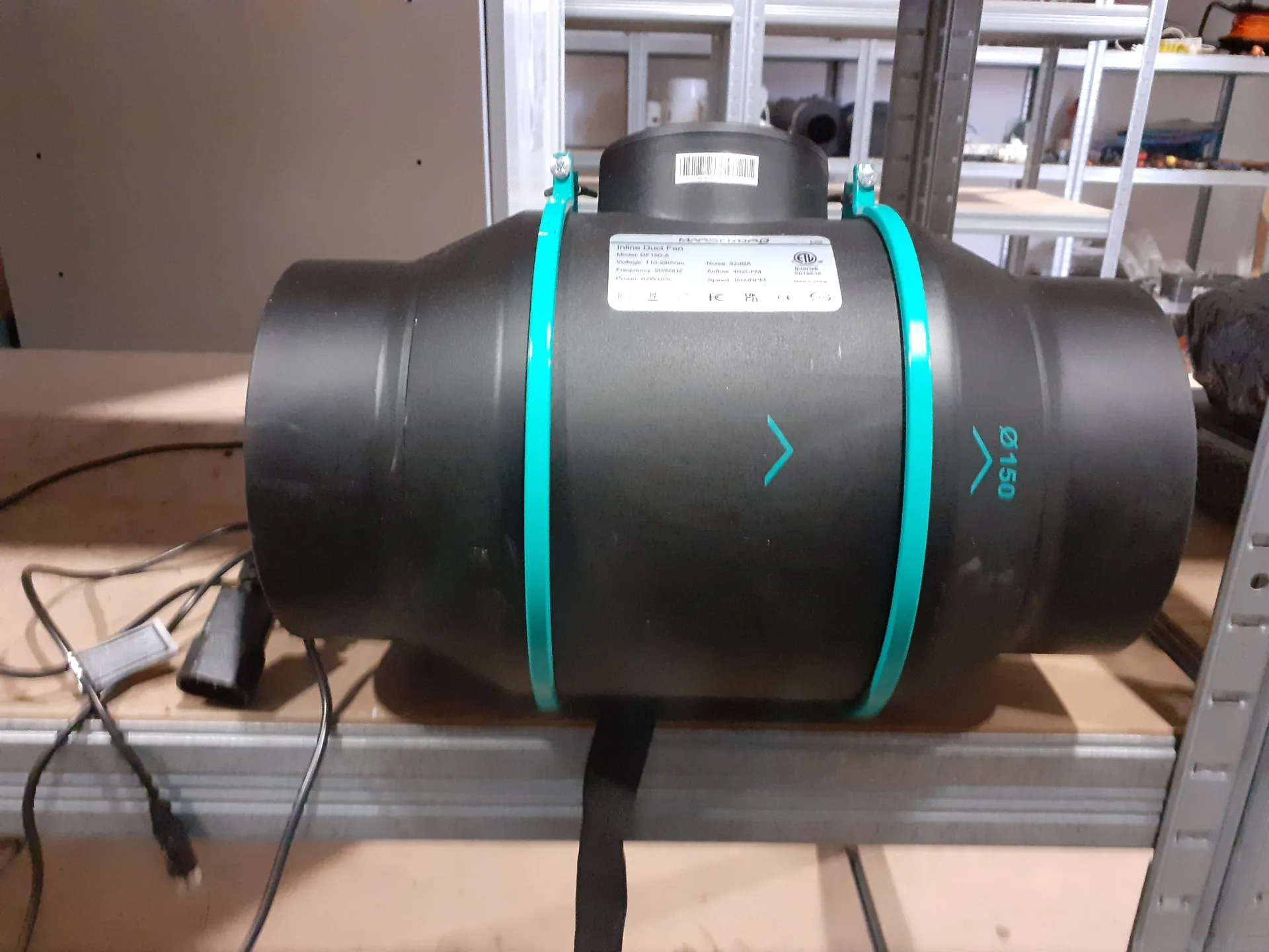 Czarny, cylindryczny wentylator kanałowy z dwiema turkusowymi opaskami stabilizującymi. Na obudowie znajduje się biała etykieta techniczna z tekstem oraz turkusowy symbol strzałki wskazujący kierunek przepływu powietrza i oznaczenie średnicy 'Ø 150'. Urządzenie leży na metalowym regale, obok widoczny jest czarny kabel z zasilaczem.