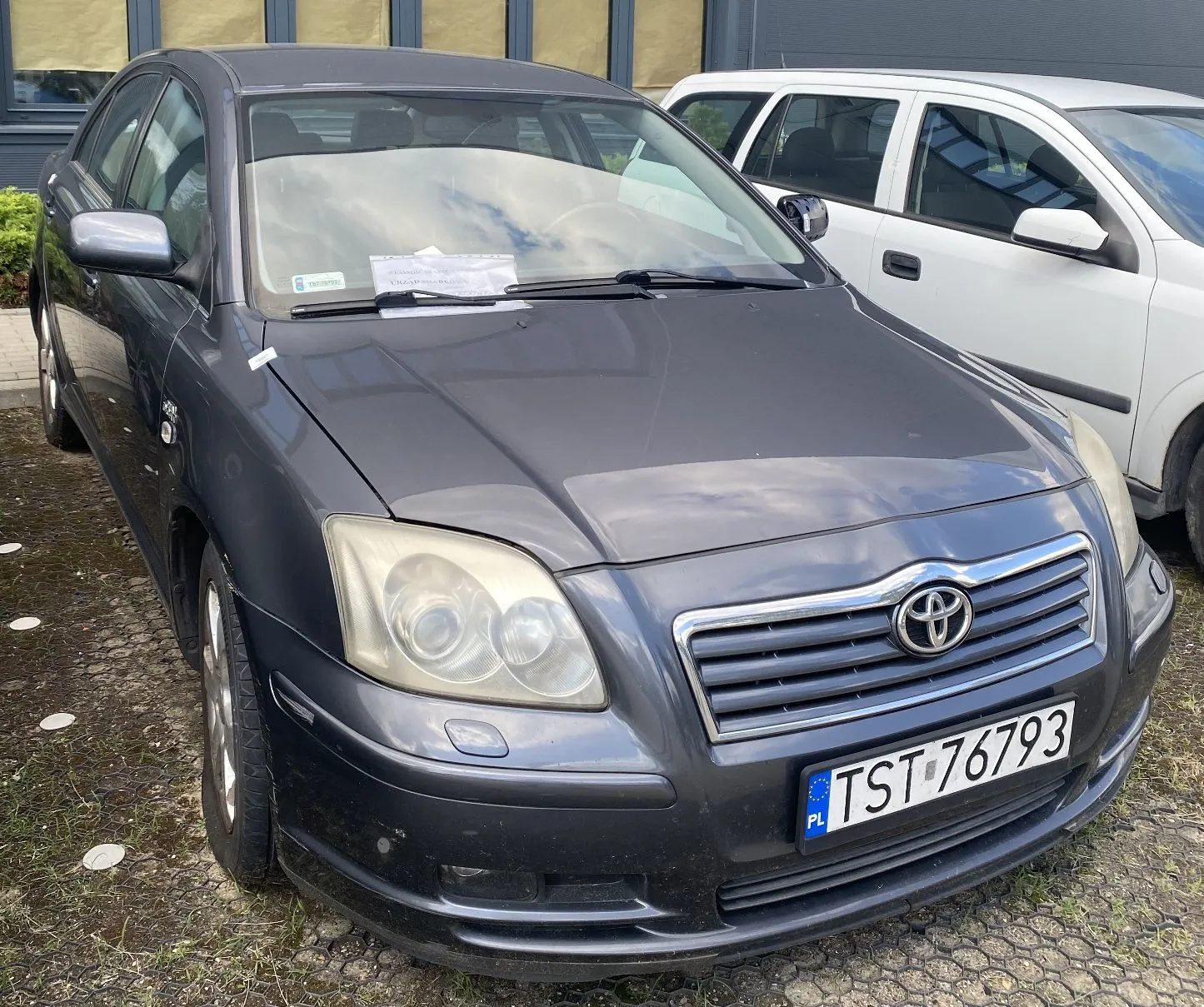 Ciemnoszary samochód osobowy marki Toyota zaparkowany obok białego samochodu. Widoczny jest przód pojazdu z logo Toyoty na grillu oraz polska tablica rejestracyjna o numerze TST 76793. Na przedniej szybie znajduje się biała kartka z tekstem.