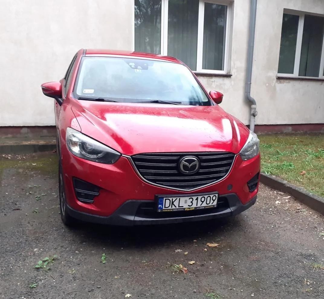 Czerwony samochód marki Mazda zaparkowany przed jasnym budynkiem. Widoczny jest przód pojazdu z polską tablicą rejestracyjną o numerze DKL 31909.