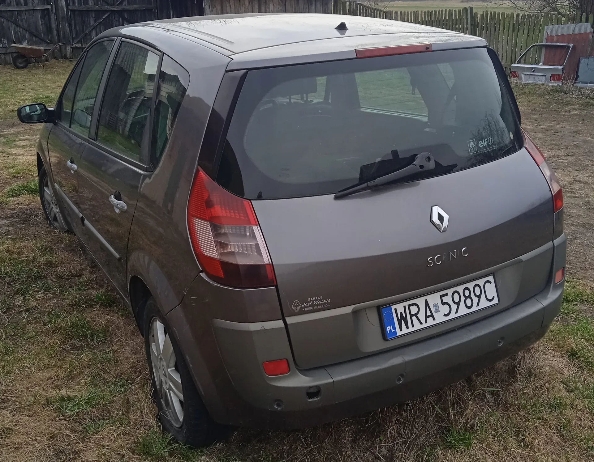 Zdjęcie przedstawia tylny widok szarego samochodu Renault Scenic zaparkowanego na trawie. Na bagażniku widoczne są logo Renault, napis 'SCENIC' oraz tablica rejestracyjna 'WRA 5989C' z niebieskim pasem 'PL'. Po lewej stronie bagażnika znajduje się naklejka z napisem 'GARAGE JOSÉ WIDMER BURG-REULAND'. W tle widoczny jest drewniany płot, sucha trawa oraz częściowo inny pojazd. Samochód ma czyste szyby i czerwone światła tylnego oświetlenia.