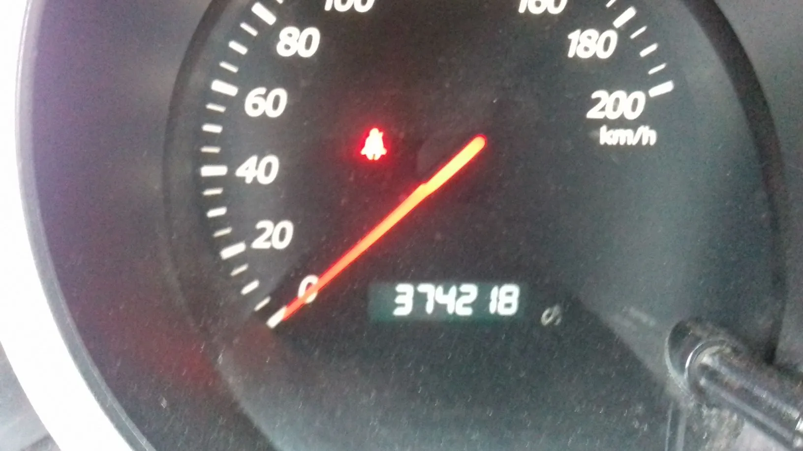 Zbliżenie na prędkościomierz samochodu. Tarcza posiada białe oznaczenia prędkości od 0 do 200 km/h. Czerwona wskazówka znajduje się w pozycji bliskiej zeru. Na środku tarczy świeci się czerwona kontrolka w kształcie dzwonka. Na dole znajduje się cyfrowy licznik przebiegu wyświetlający liczbę 374218.