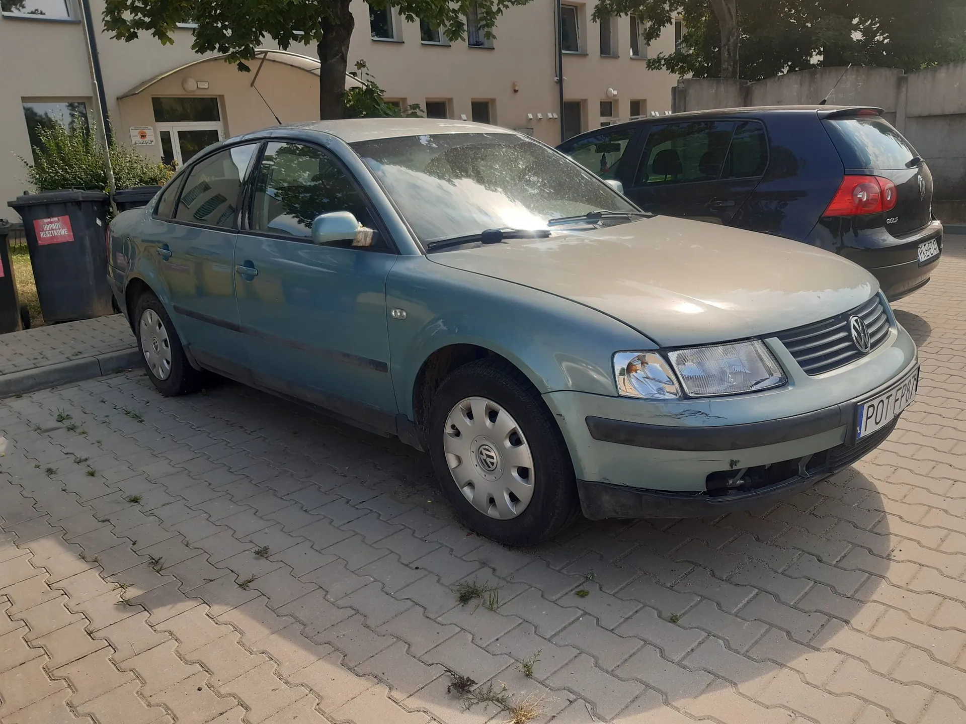 Jasnoniebieski samochód osobowy marki Volkswagen zaparkowany na kostce brukowej przed jasnym budynkiem. Obok stoi ciemny samochód typu kombi. W tle widoczne są drzewa i fragmenty elewacji budynku.
