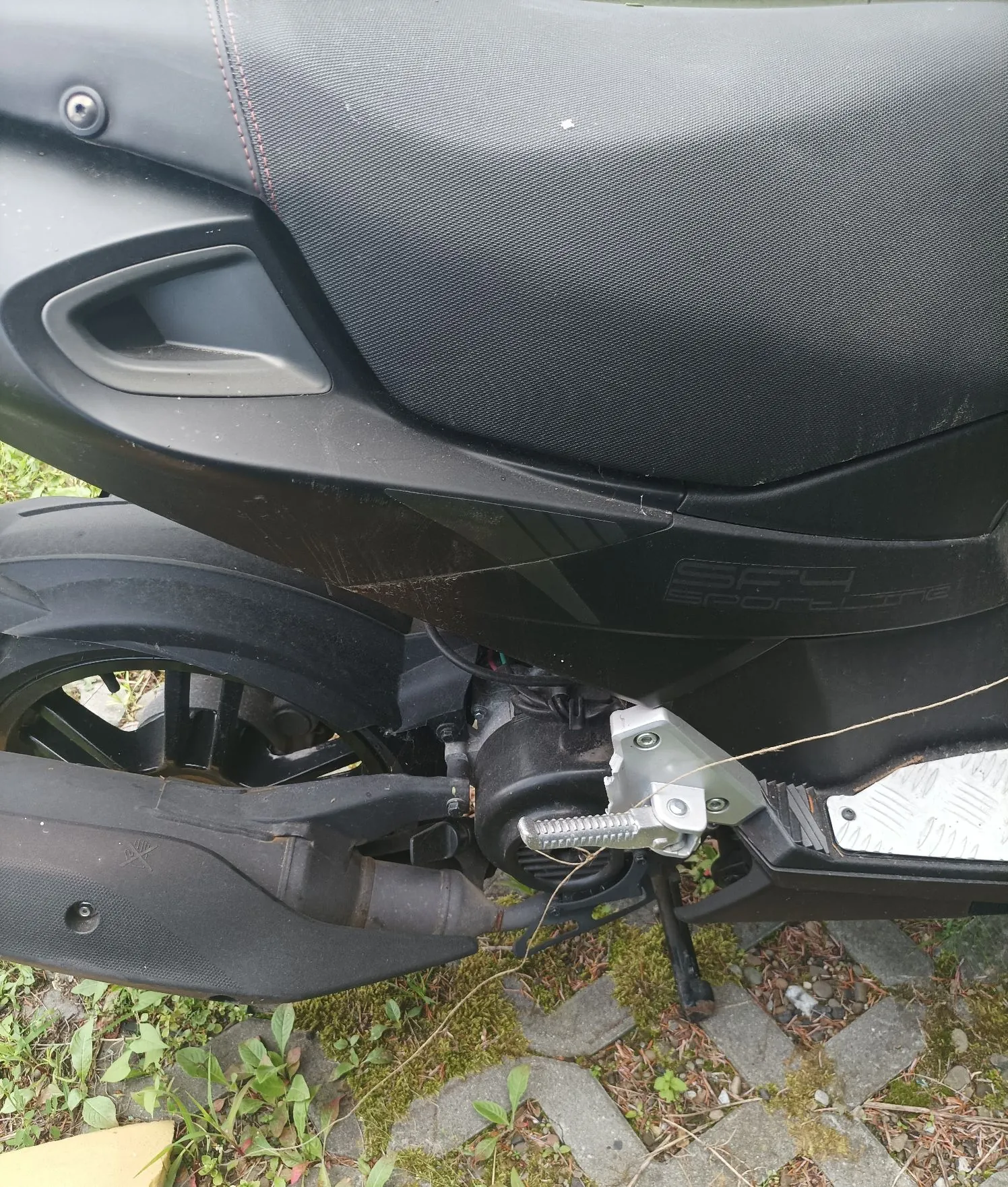 Zbliżenie na tylną część czarnego skutera. Widoczny jest fragment siedzenia z czerwoną przeszyciem, czarna obudowa z napisem 'SF4 SPORTING', tylne koło, czarny wydech oraz metalowa stopka. Skuter stoi na kostce brukowej z przerastającą trawą.