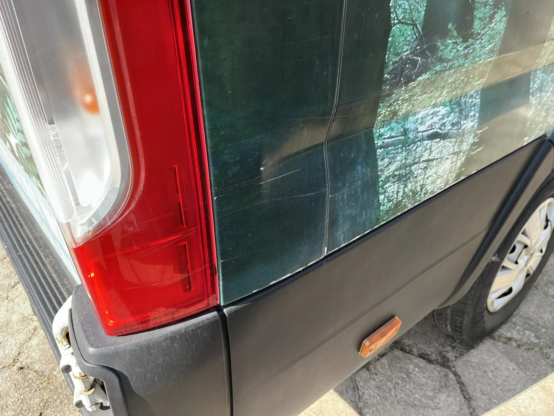 Zbliżenie na tył ciemnozielonego pojazdu. Widoczna jest czerwona lampa tylna oraz czarny plastikowy zderzak. Na karoserii nad zderzakiem znajdują się liczne zarysowania oraz wgniecenie. W lakierze odbijają się drzewa i roślinność.